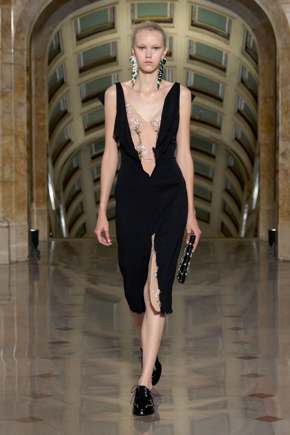 Black crepe dress with plunging raw-edged panels framing nude floral tulle. Mismatched blossom-drop earrings in white, black, and green stones. Slim black clutch dotted with tiny white florets. Glossy black derbies with neat round laces. Crossing a museum atrium after midnight rain, flowers crushed in one hand.