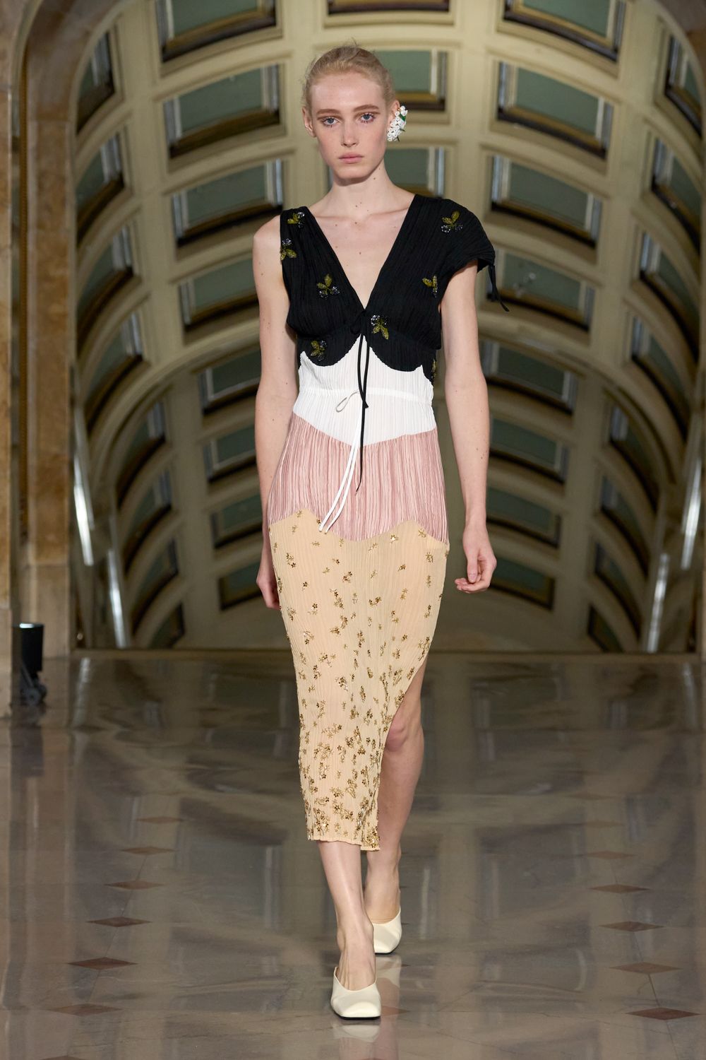 Crinkled slip dress color-blocked in white, shell pink and buttercream, scattered with tiny gold sprigs. Black pleated capelet with beaded leaf clusters and one loose tie end. Cream leather pumps with squared toes. Crossing a marble atrium after the wedding guests have gone upstairs.