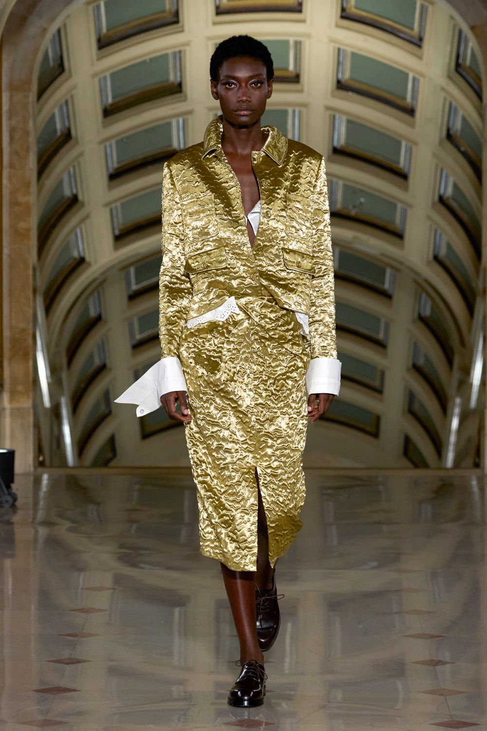 Antique-gold moire jacket with broad flap pockets and stark white cuffs. White triangle bra peeking through the deep open placket. Matching column skirt, white eyelet pocket linings pushed out at the hips. Dark oxblood derbies with thin laces. Crossing a civic rotunda after the wedding, still carrying the tablecloth sheen.