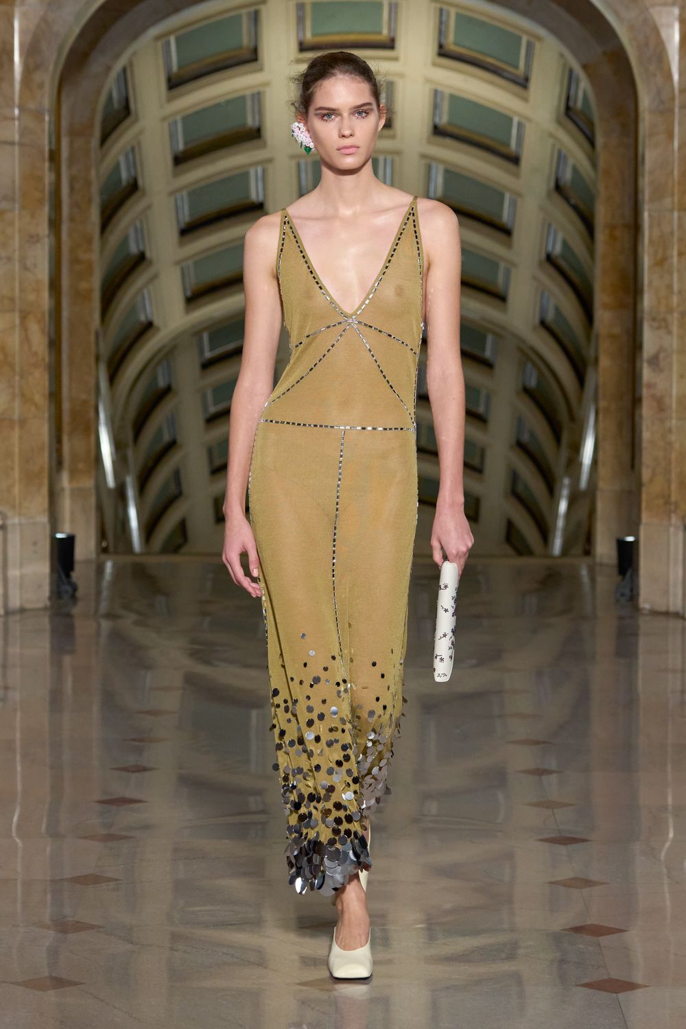 Clustered pale blossom pinned against one ear. Moss knit slip dress, mirror-strip seams and paillette-heavy hem. Narrow ivory cylinder clutch with tiny black motifs. Cream pumps with a squared toe. Crossing a museum atrium just after the string quartet packed up.