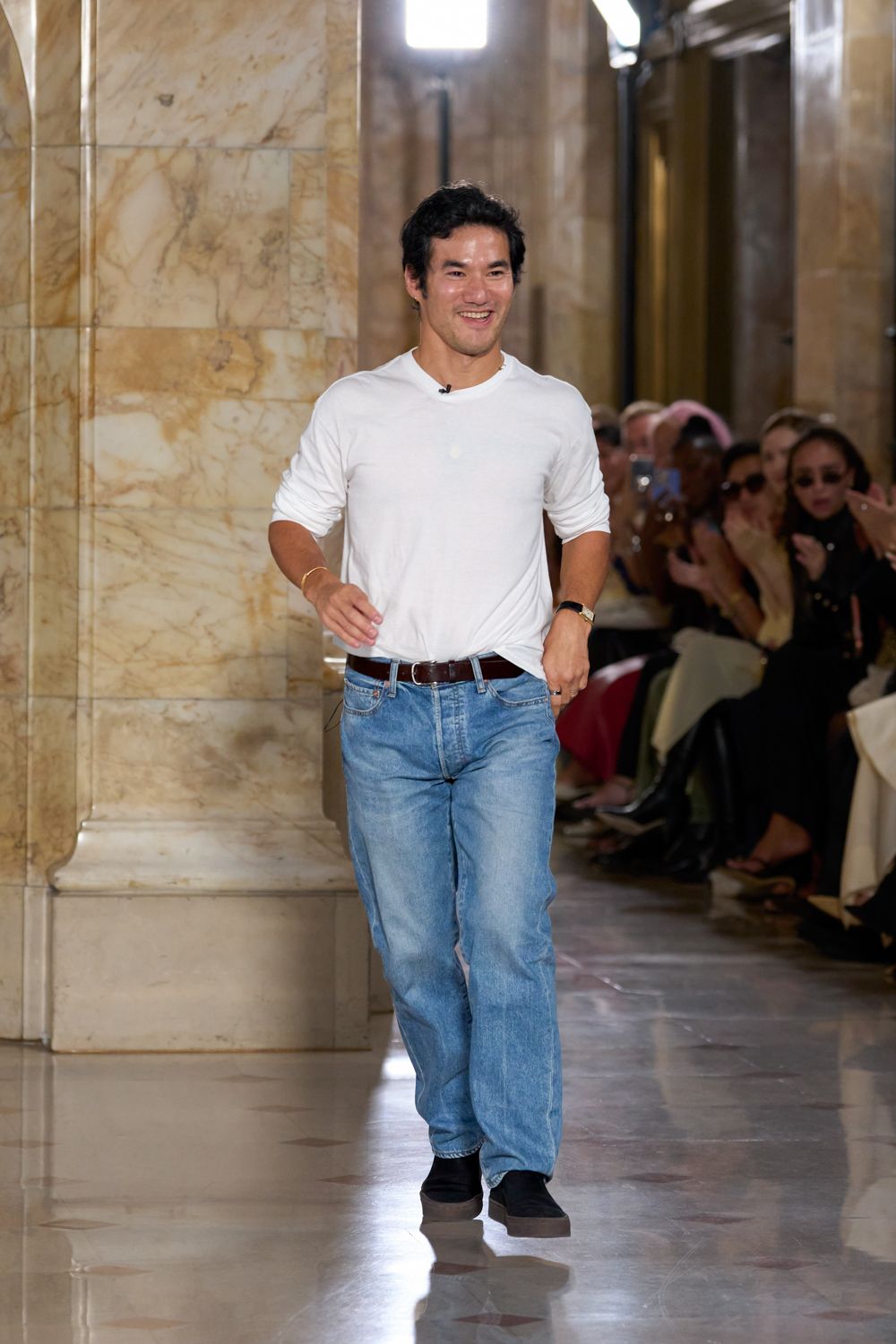 White jersey long-sleeve tee, sleeves shoved up and hem loosely tucked. Light-wash straight jeans with whiskering and a soft fade through the thigh. Dark brown leather belt with a small silver buckle. Black slip-ons on thick gum soles. Walking across a marble lobby after everyone else has already gone upstairs.