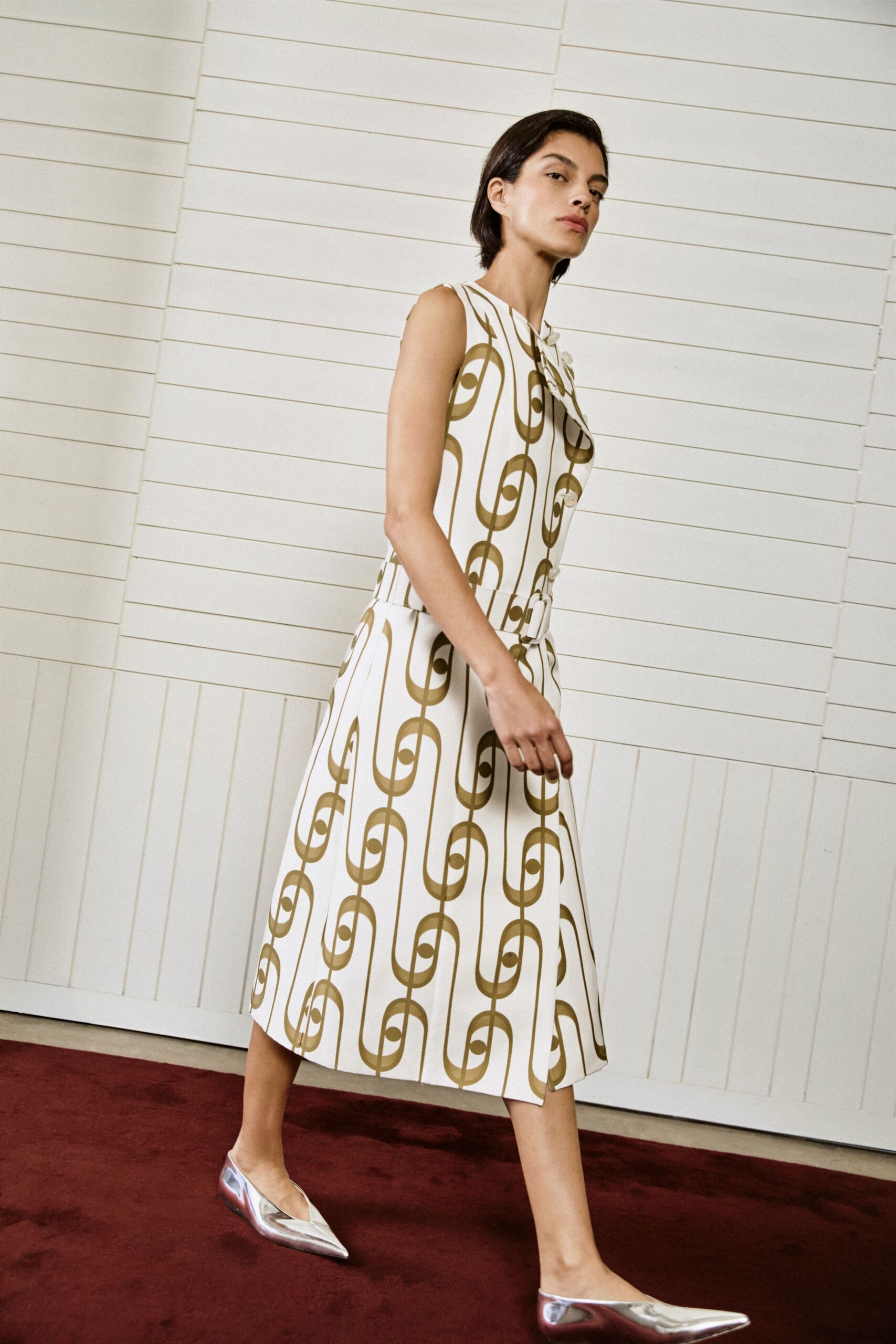 Ivory sleeveless dress in stiff twill, olive chain print and belted waist. Mirror-silver pointed flats catching red from the carpet. Walking a quiet hotel corridor before the luncheon doors open.