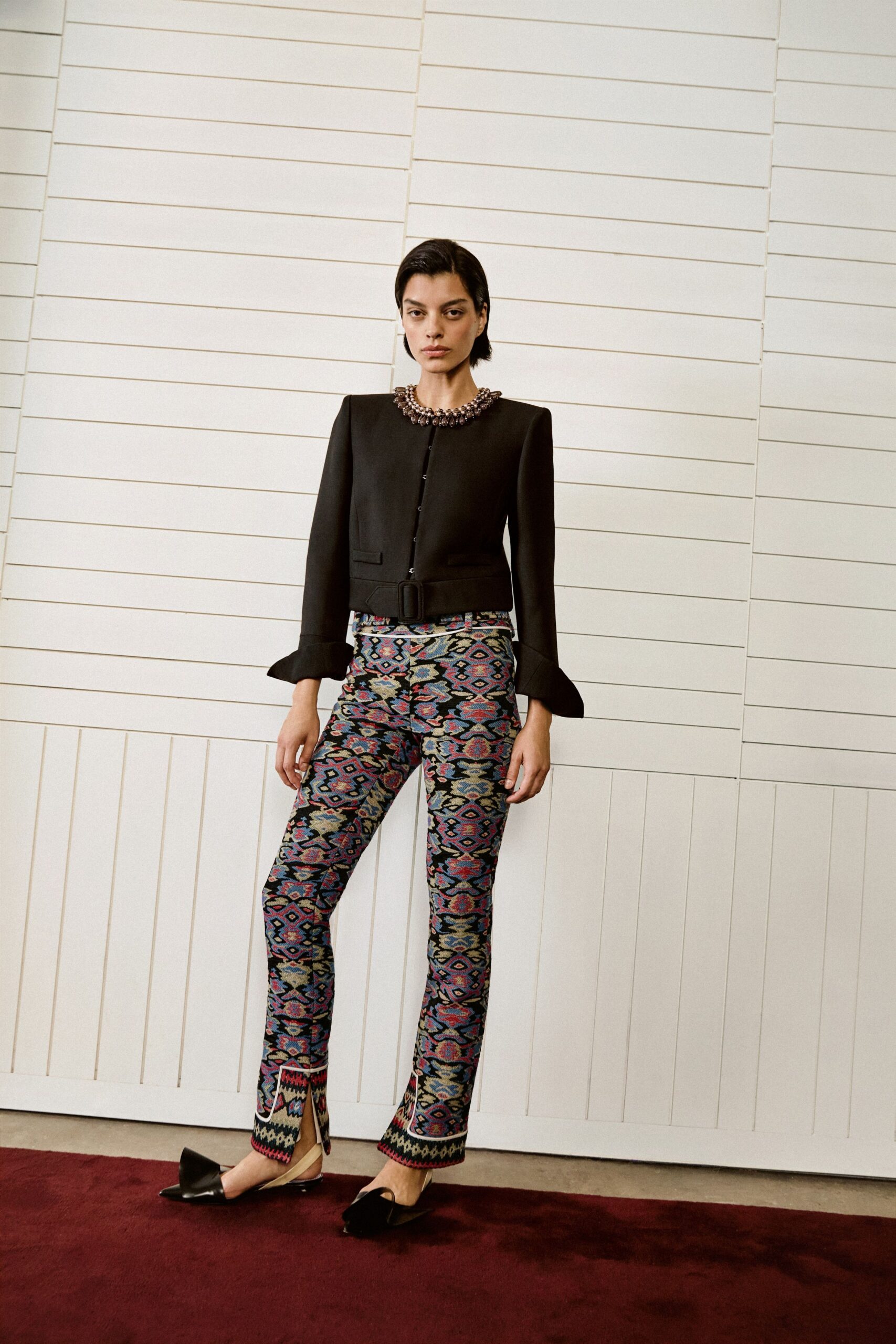 Black cropped jacket with a jeweled collar and flared turned-back cuffs. Wide self belt fastened with a square covered buckle. Slim jacquard trousers in a carpet-like ikat, split at the front hem. Black satin slingback flats with oversized folded bows. For crossing a hotel corridor before the wedding guests start arriving.