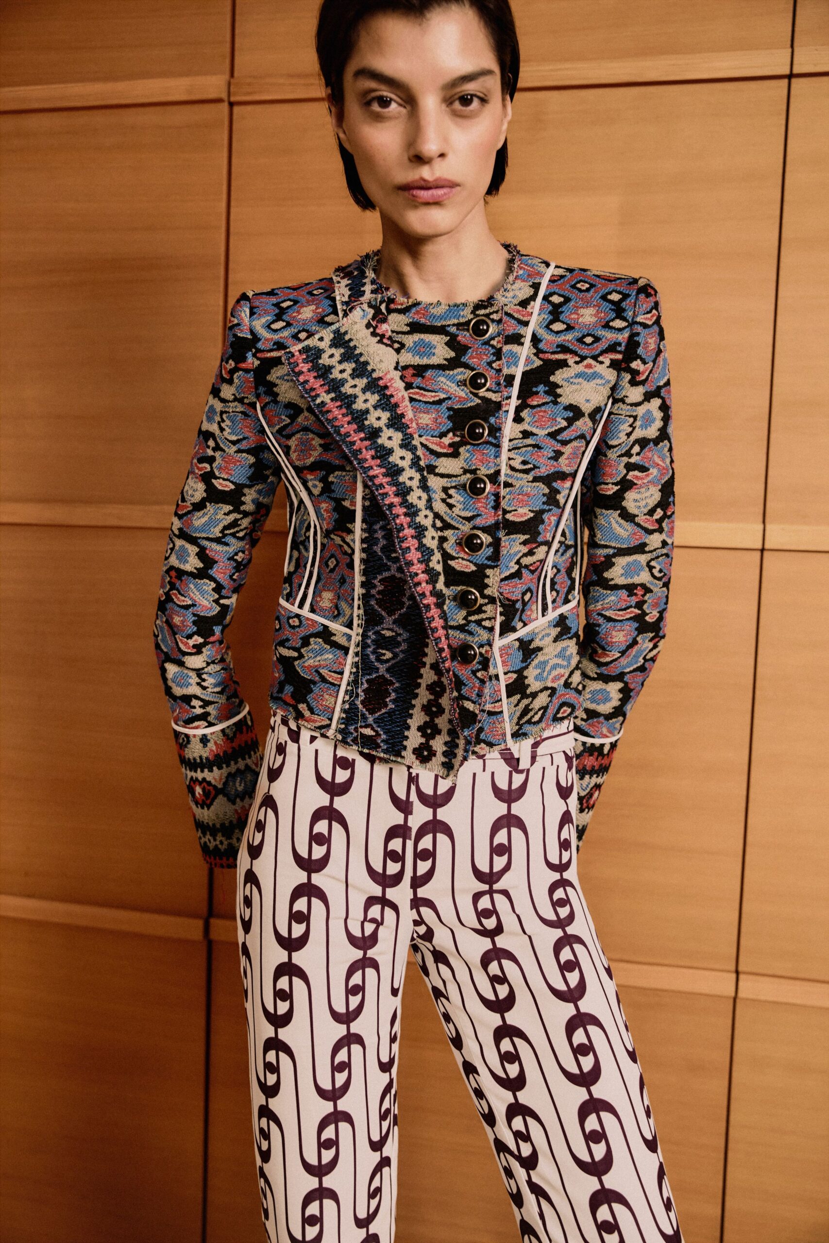 Fitted tapestry jacket in blue and cream jacquard, white piping tracing every seam. Diagonal woven panel with pink blanket-stitch edges crossing the button front. White flared trousers printed with plum chain-link stripes. Hotel corridor paneling, waiting for the private dining room to open.