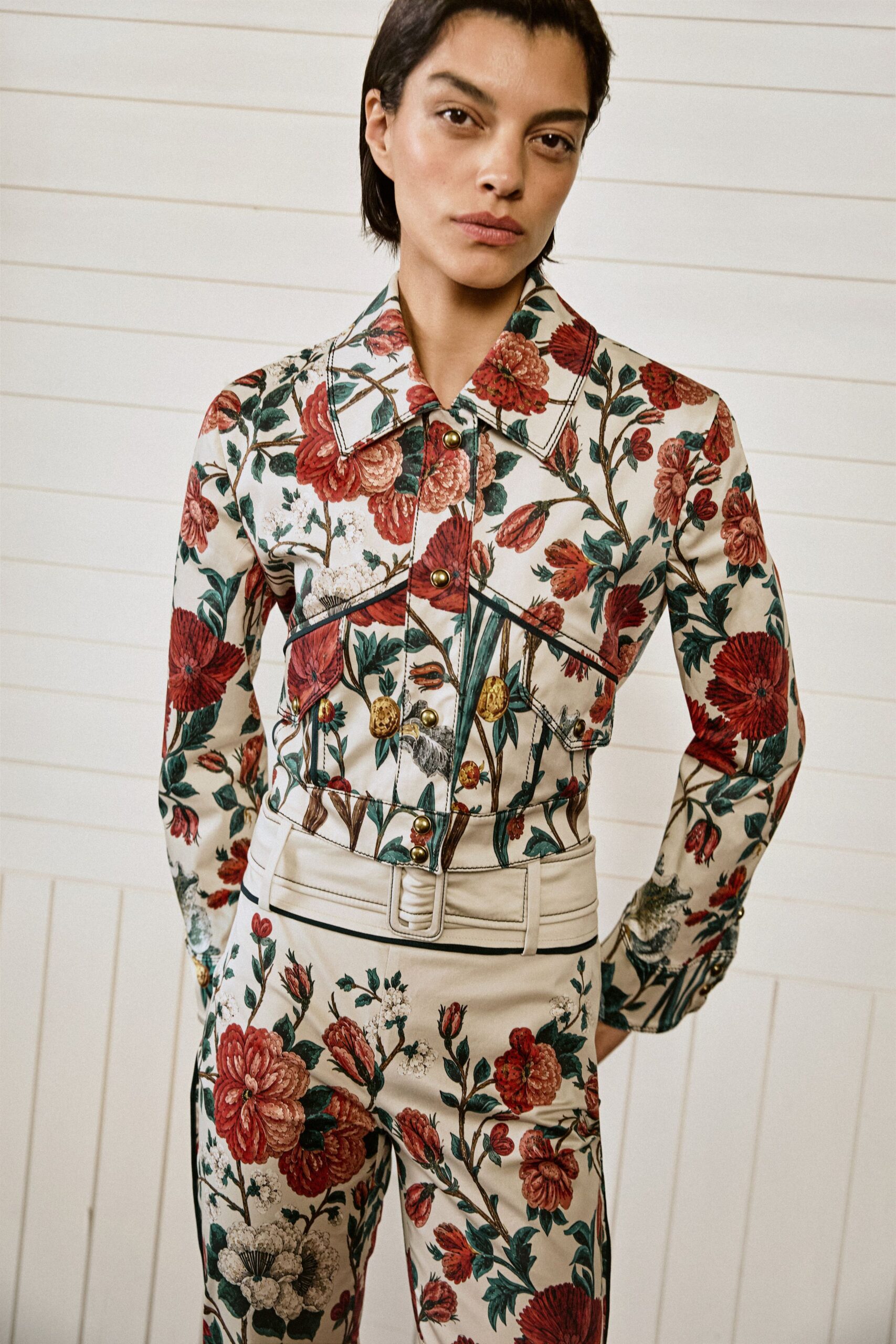 Cream floral jacket in smooth twill, black piping tracing the pointed yoke. Matching high-waisted trousers with a wide self belt and padded buckle. For walking through a conservatory after the rain, shoes clicking on damp tile.