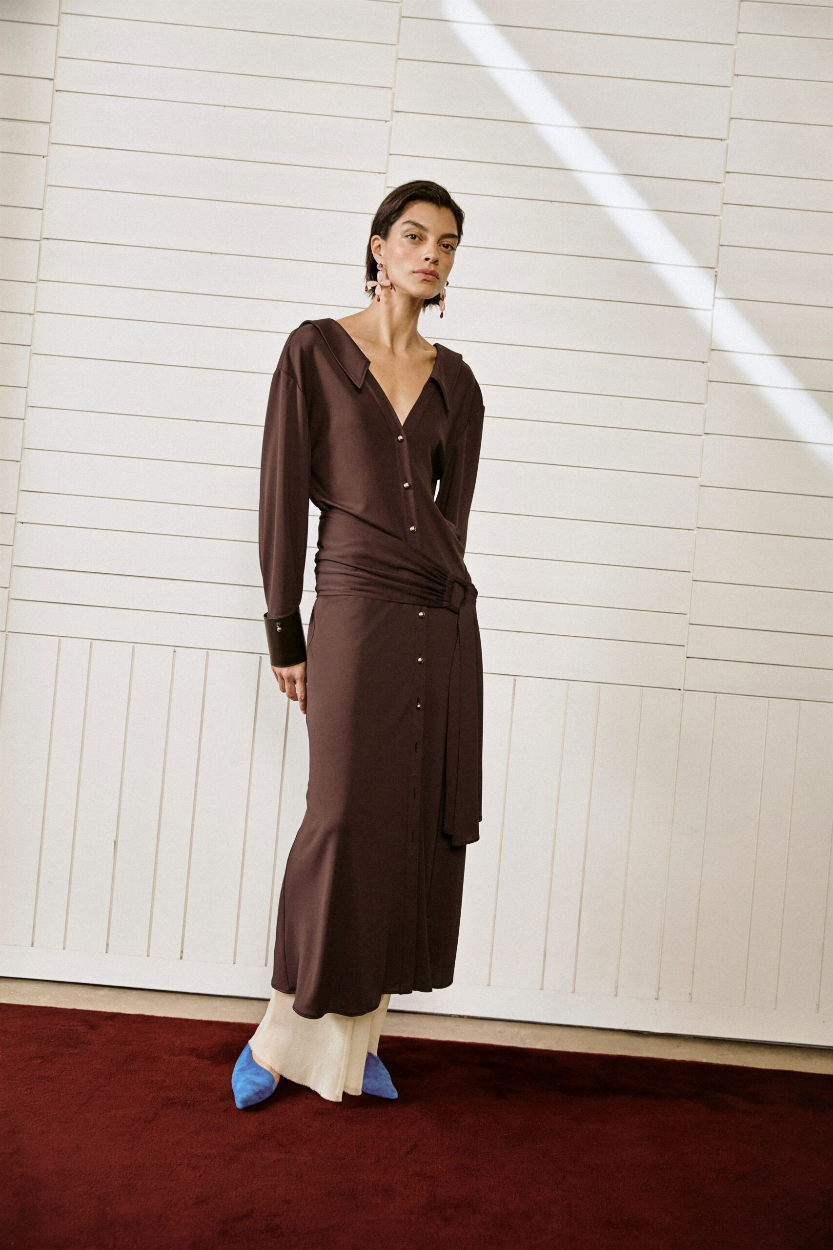 Blush petal-drop earrings brushing the jaw. Espresso jersey shirt dress, silver buttons and a ruched sash crossing low. Cream wide-leg trousers pooling below the hem. Cobalt suede pointed flats. Afternoon check-in at a seaside hotel, sun striping the lobby wall.