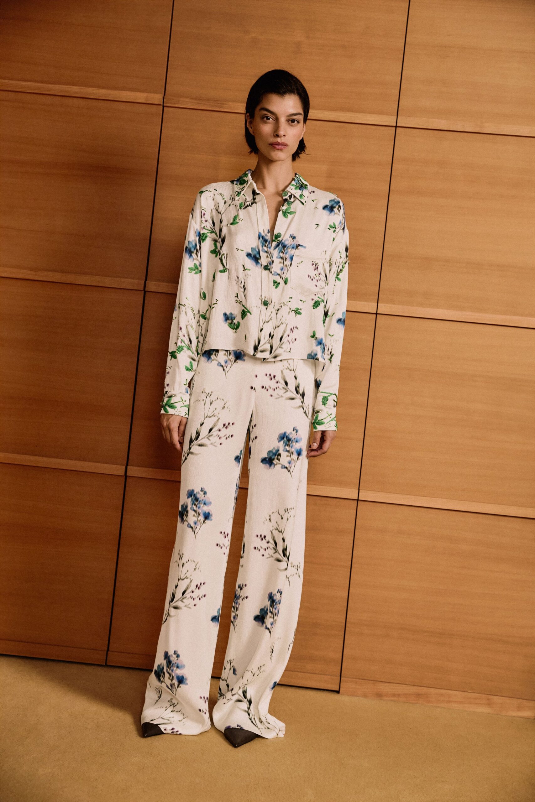 Ivory crepe blouse scattered with blue wildflowers, one flap hanging loose at the placket. Matching wide-leg crepe trousers, pooling over the shoes in the same print. Black pointed pumps barely breaking the hem. Hotel corridor at noon, flowers from yesterday still holding up.