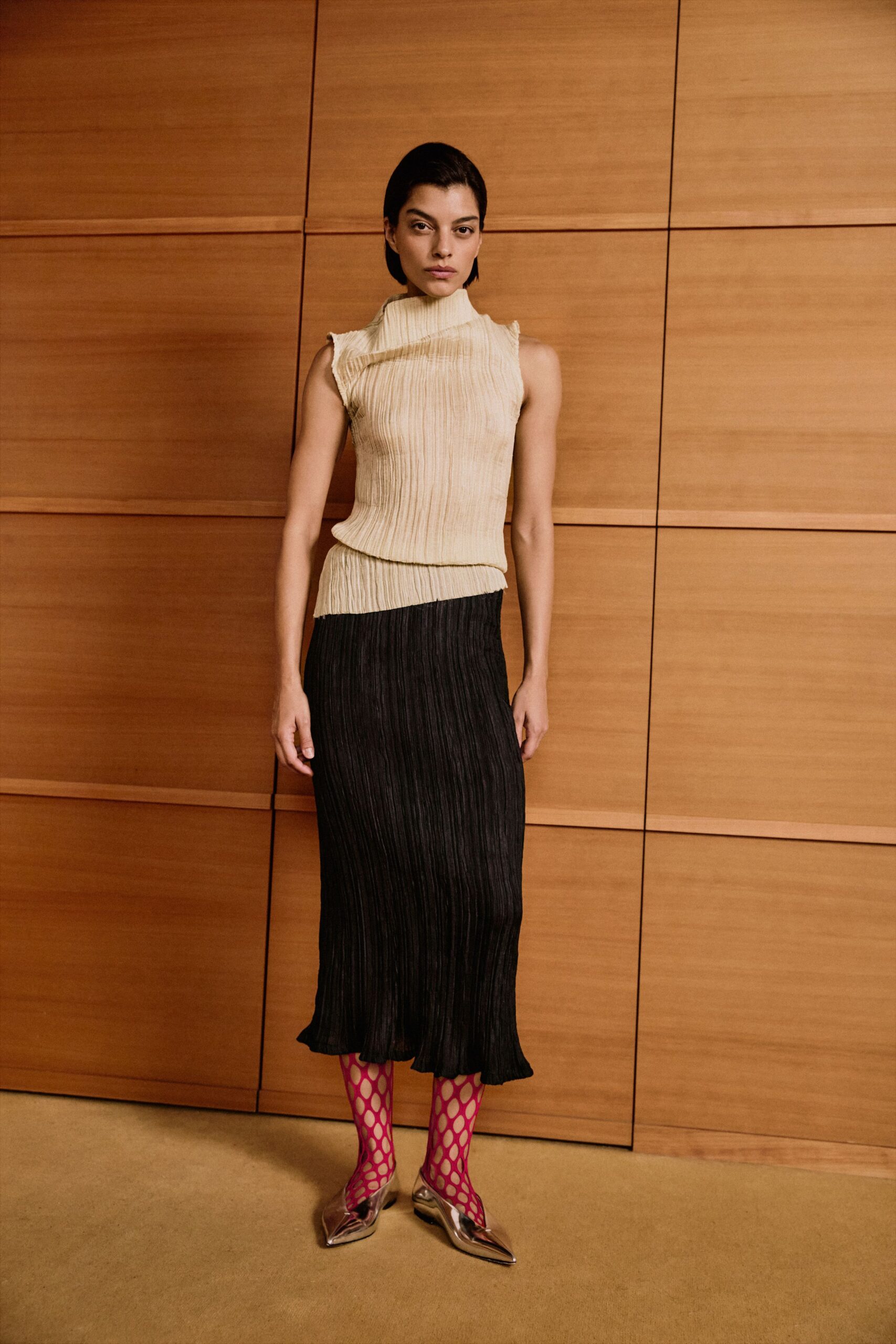 Cream crinkled knit shell with an off-kilter funnel neck and raw armholes. Black crinkled column skirt, waistband sliced on the diagonal. Hot-pink diamond fishnet tights. Mirror-silver pointed flats, toes bent upward. Crossing a hotel atrium before the conference coffee has been cleared.