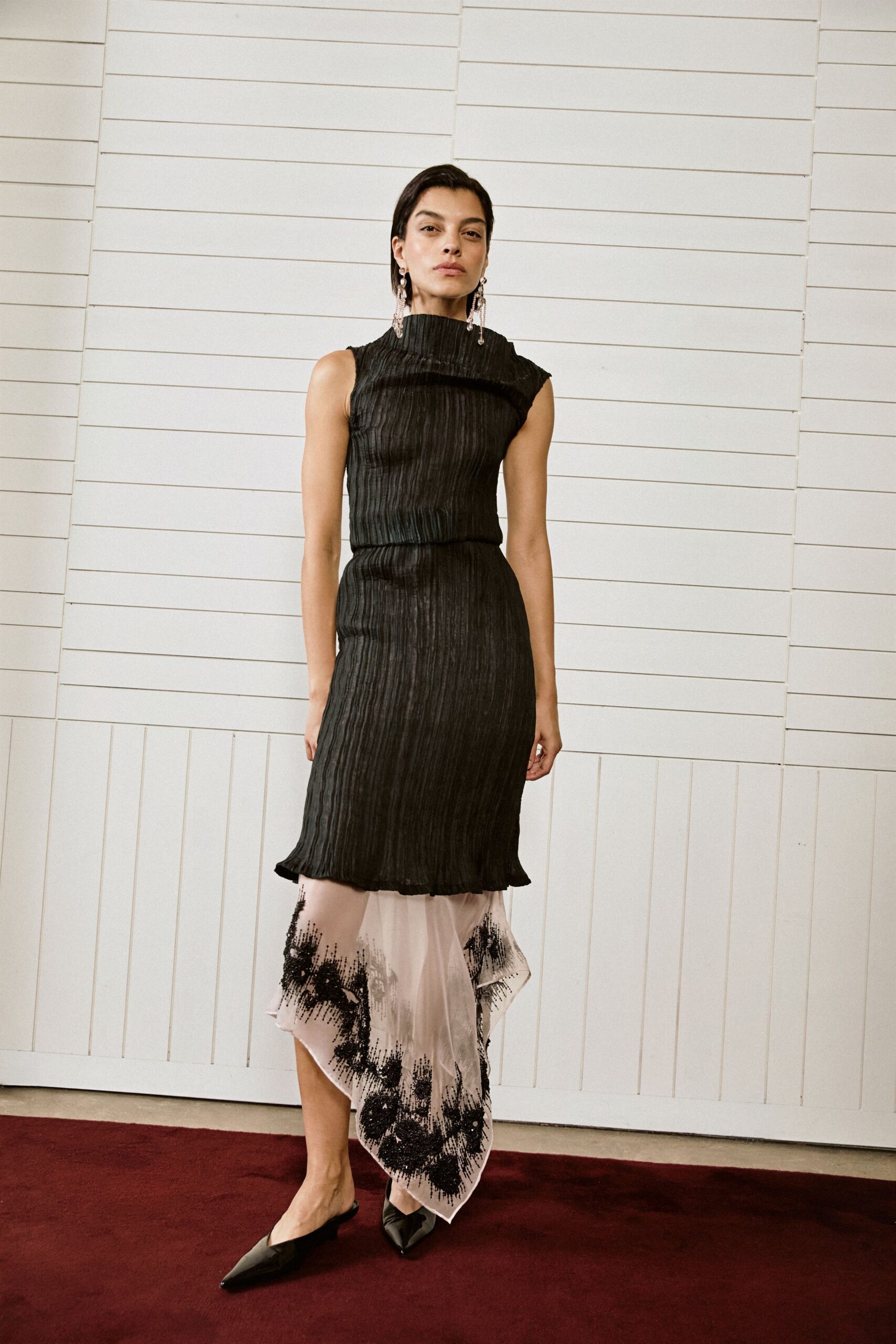 Black crinkled plisse dress with a rolled funnel neck and dropped overlay. Blush sheer underlayer falling past the hem in black beaded bursts. Long crystal drop earrings. Black pointed mules. Crossing a hotel corridor before the wedding crowd spills out.
