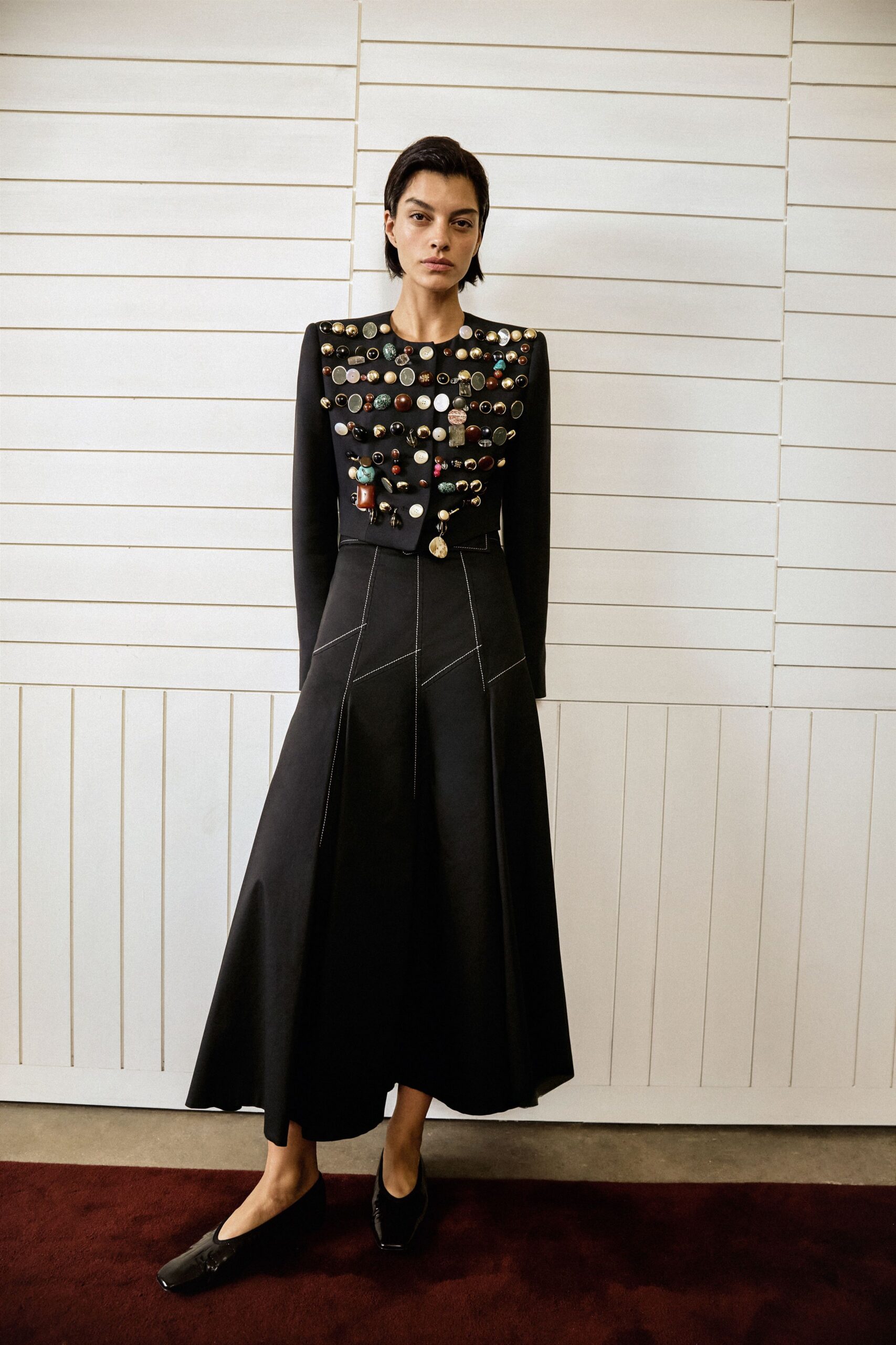 Cropped black jacket crusted with mismatched brooches, charms and cabochons. Black flared midi skirt with white topstitching tracing angled seam panels. Square-toe black flats, glossy at the vamp. House museum corridor, pausing under portraits that all seem to disapprove.