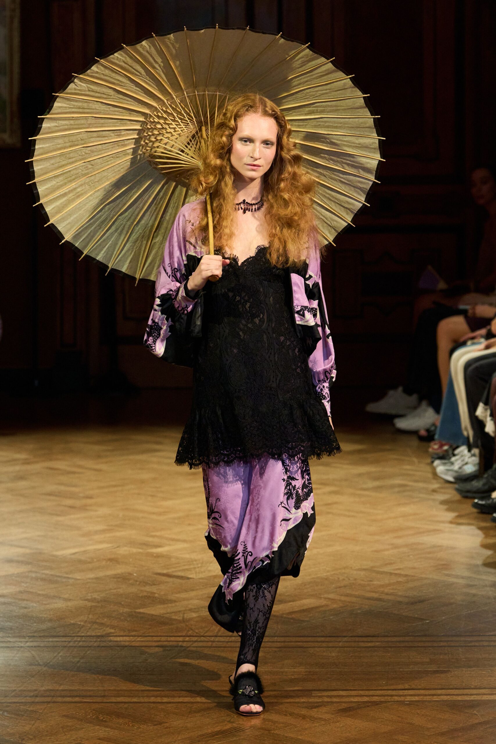 Pale paper parasol with bamboo spokes fanning behind the head. Lilac satin robe printed with black florals and wide sleeves. Black lace slip dress, scalloped hem falling over the robe. Short jet-bead necklace. Black floral lace tights. Black mules with fluffy vamp trim. Crossing a drafty hotel corridor after midnight rain.