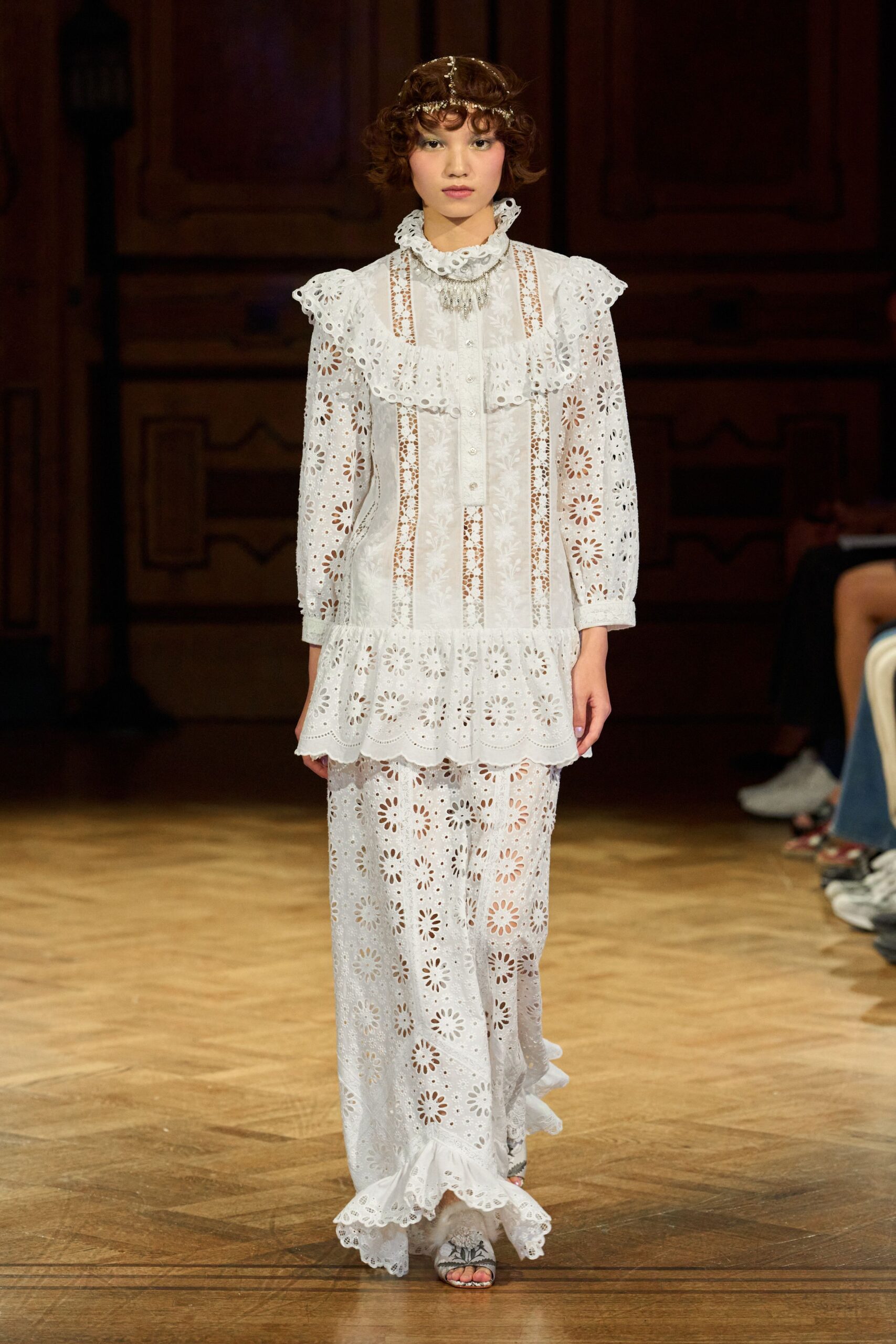 Fine silver chain headpiece draped over short finger-wave curls. White broderie anglaise dress with ruffled collar, bib buttons, and dropped flounce hem. Matching eyelet trousers, wide through the leg with a ruffled cuff. Pale floral sandals peeking under the hem. Afternoon tea in a drafty manor, rain tapping the tall windows.