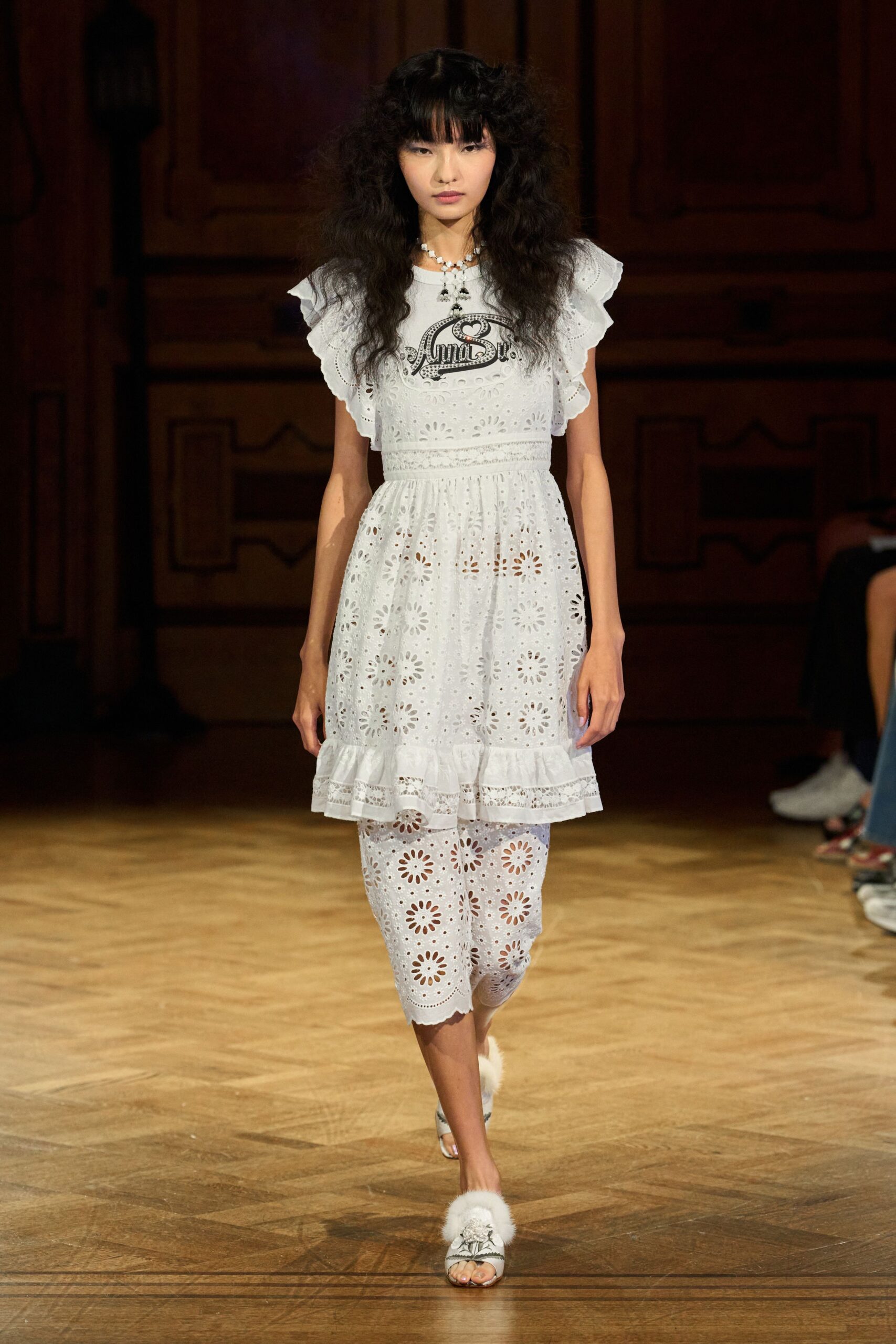White broderie anglaise dress with flutter sleeves and a lace-set dropped waist. Matching eyelet capris, scalloped just below the knee. Clustered pearl charms at the collarbone. White open-toe mules topped with fluffy marabou puffs. Sunday hotel breakfast, still in yesterday's hair.