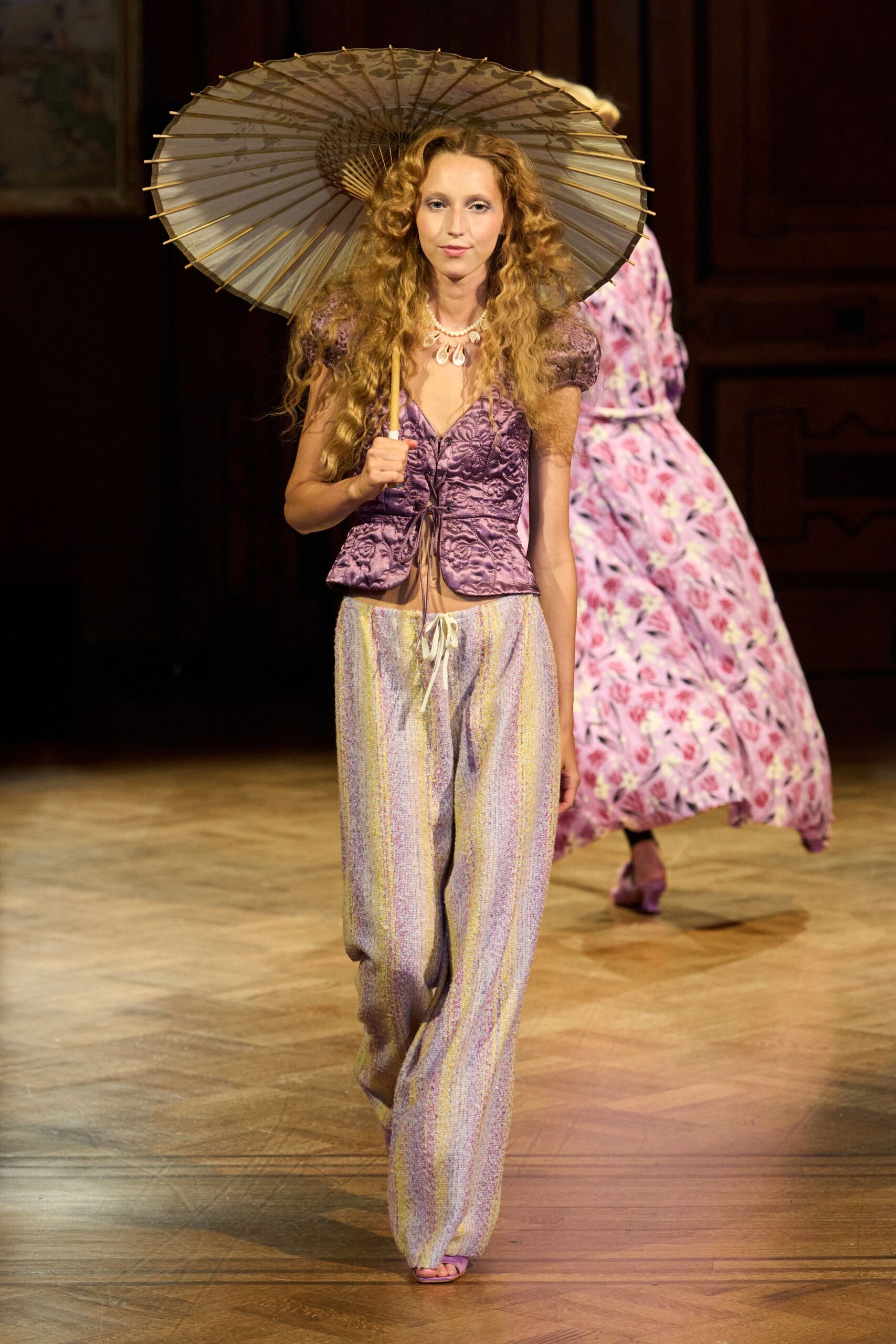 Pale paper parasol with bamboo spokes radiating behind the head. Short pearl strand with shell drops at the collarbone. Mauve brocade blouse, puff sleeves and dangling front tie. Loose pastel bouclé trousers with a white drawstring knot. Lilac open-toe mules. Crossing the lawn after a garden wedding, cake plate still in hand.