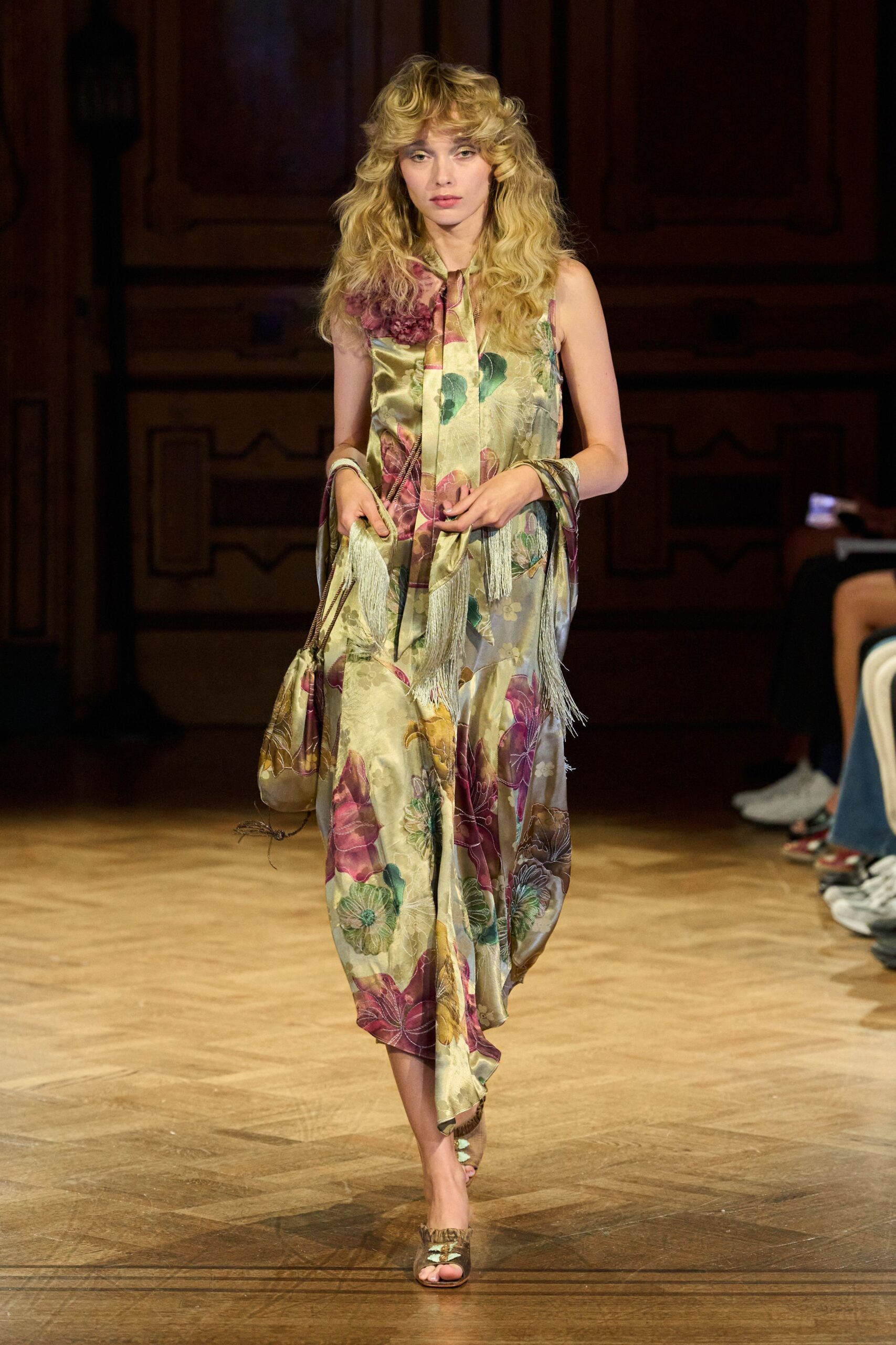 Sage satin slip dress printed with oversized mauve blooms, scarf panels swinging fringe. Matching satin drawstring pouch, cord end trailing loose. Olive satin mules with ruffled fringe across the toes. Crossing a hotel ballroom after the wedding band has packed up.