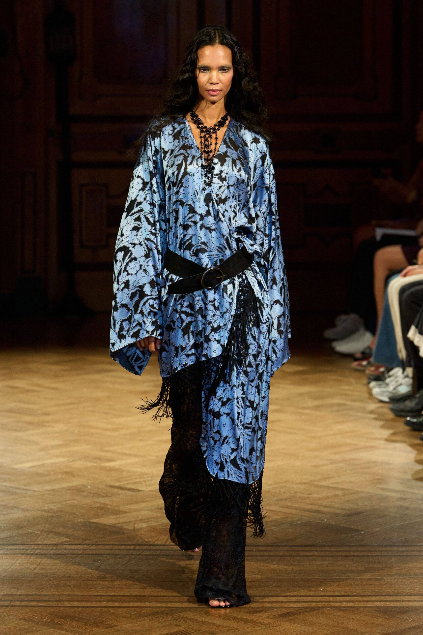 Tiered black bead necklace dropping nearly to the waist. Powder-blue floral satin caftan, one side draped long with fringe. Wide black belt cinched low with a round buckle. Sheer black lace trousers, pooling over the toes. Crossing a hotel ballroom after midnight, carrying your shoes by hand.