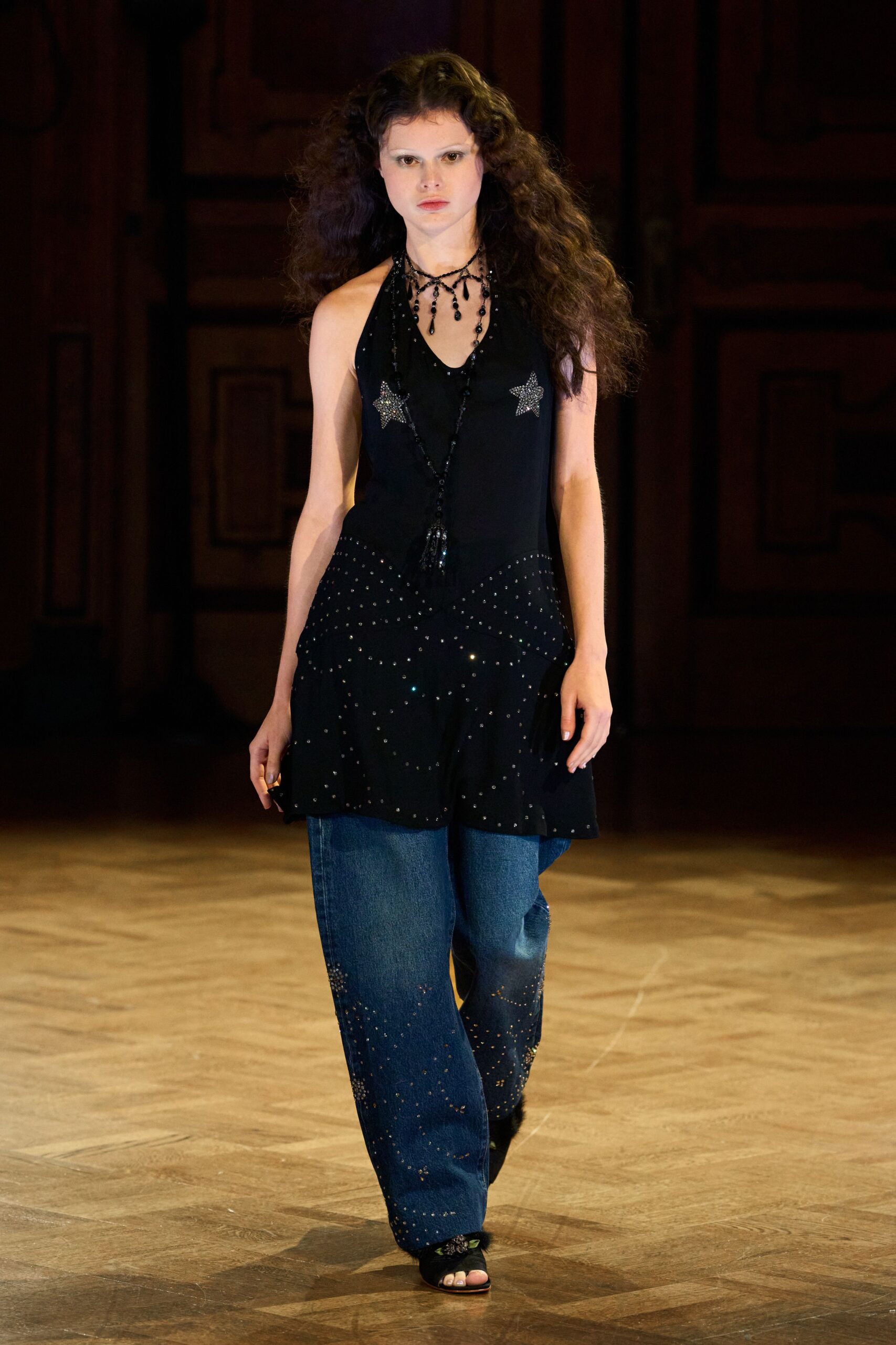 Black beaded bib necklace dripping teardrop pendants and a long tassel. Black halter tunic with rhinestone stars and a studded flounce hem. Wide indigo jeans scattered with tiny crystals, densest near the cuffs. Black open-toe mules with a glittering floral vamp. After the county fair, still dressed for the song you sang.