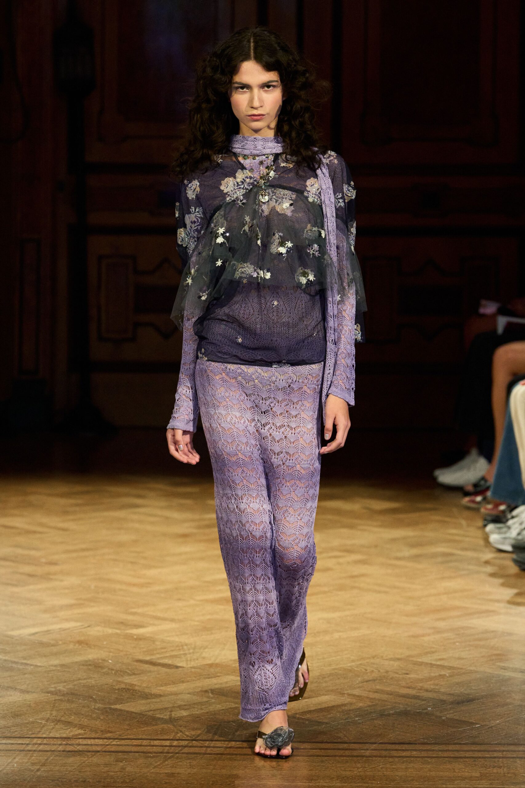 Lavender lace scarf looped close, one end falling past the waist. Sheer violet mesh top scattered with faded blossom embroidery. Lilac openwork lace trousers, the hem pooling lightly over the foot. Black thong sandals with a crumpled flower at the toe. Crossing a hotel ballroom after the wedding band has packed up.