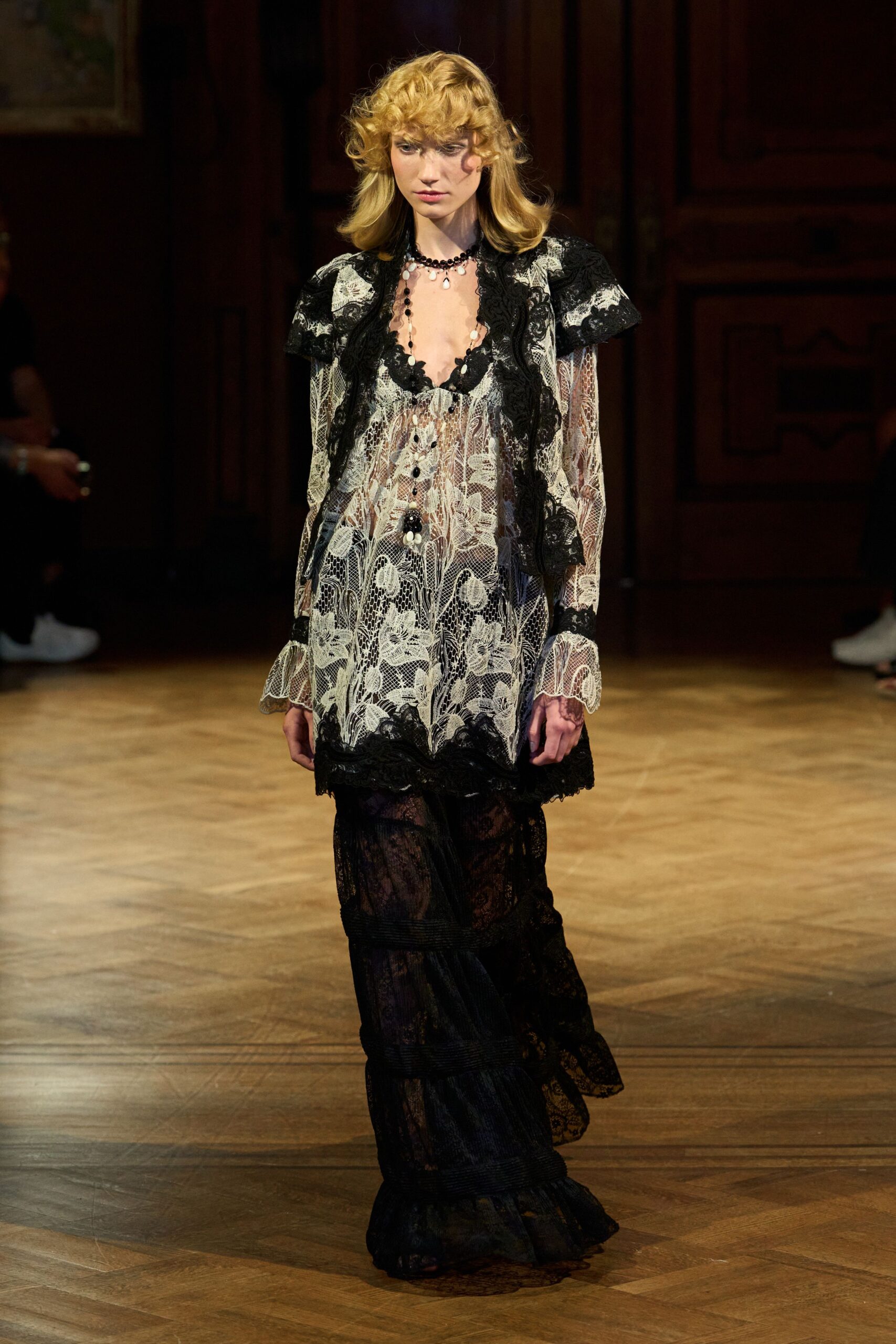 Black beads with milky teardrop charms, one dangling to the sternum. Ivory floral lace slip dress with scalloped black trim and bell cuffs. Sheer black lace maxi underlayer, tiered with narrow ribbed bands. Afternoon wedding in a drafty manor, rain starting against the windows.