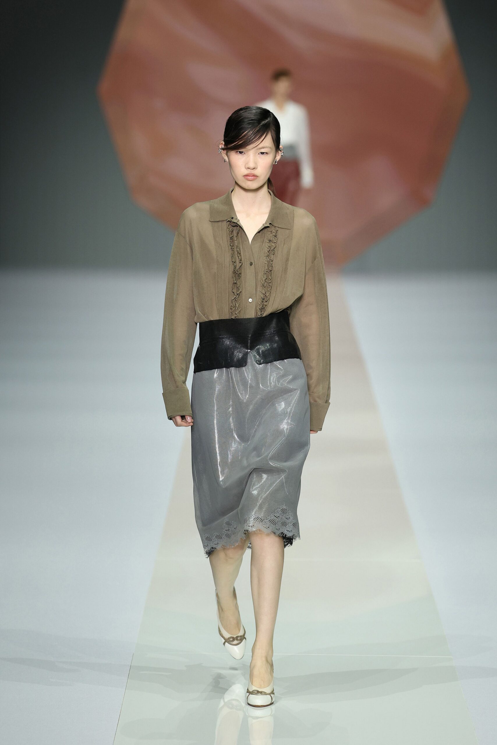 Taupe sheer blouse with twin ruffled plackets and slouched cuffs. Glossy black leather cincher cut in a deep curved point. Pewter lacquered skirt, eyelet lace peeking from the uneven hem. Cream pointed flats with a chain bit across the vamp. Hotel lobby after rain, still carrying the conference badge in your pocket.