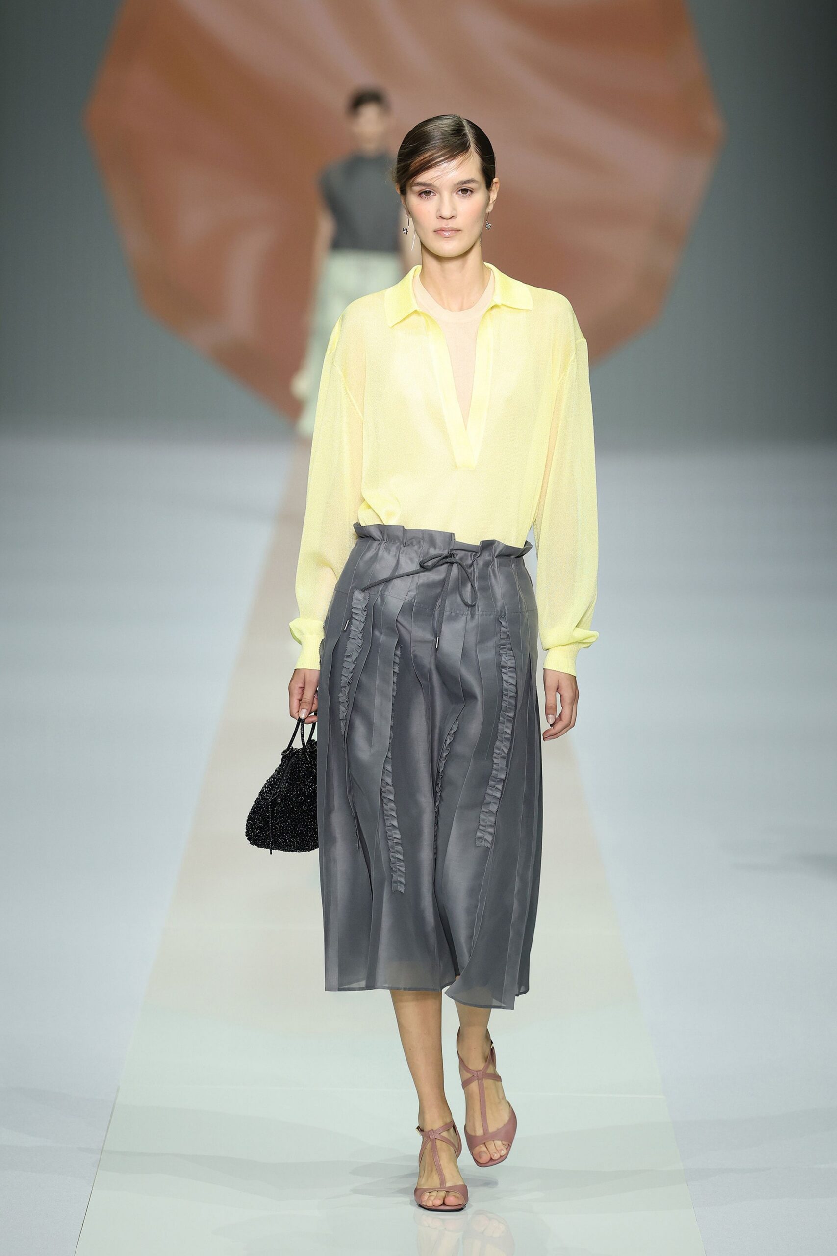 Pale citron sheer blouse with a deep split placket. Smoke-grey midi skirt, paperbag waist tied with a limp cord. Black beaded drawstring pouch. Dusty mauve T-strap heels. Walking back from a hotel terrace lunch, air still heavy with rain.