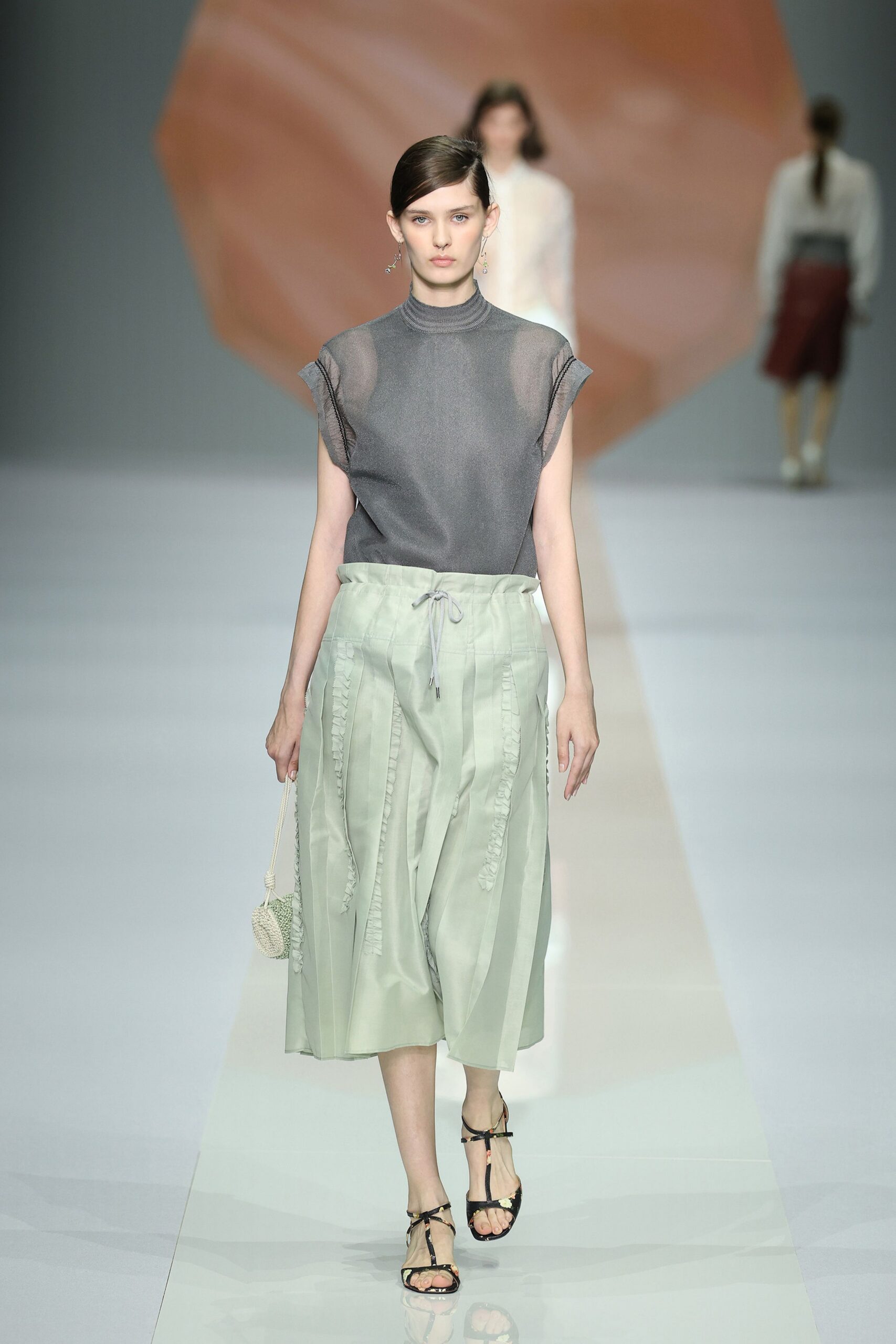 Smoky grey sheer knit top with rolled cap sleeves and mock neck. Pale celadon culottes, softly striped with vertical ruffled seams and drawstring waist. Tiny knitted pouch on a rope wrist loop. Black T-strap sandals with slim floral-printed straps. Crossing a museum atrium with cut flowers still wrapped in paper.