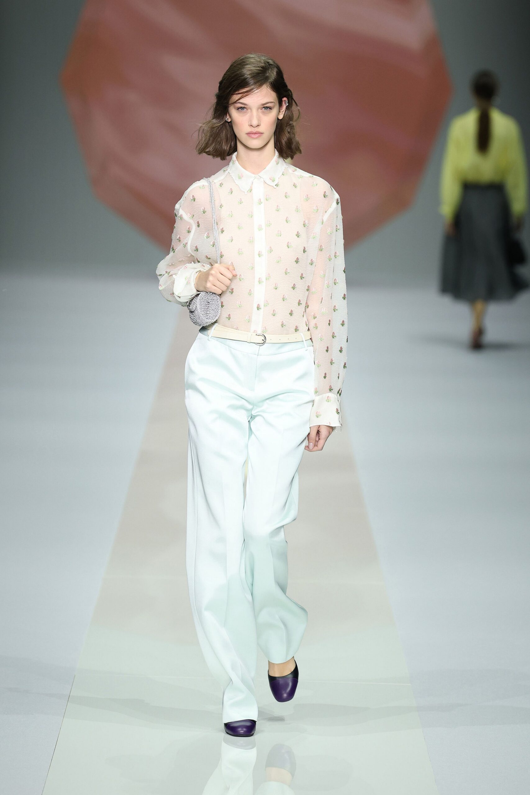 Sheer blush blouse scattered with tiny sprig motifs and a scalloped collar. Silver beaded pouch hanging soft from a chain strap. Narrow white belt with a small ring buckle. Chalk wide-leg trousers, pressed flat through the front. Deep violet cap-toe pumps. Walking into a hotel atrium with peonies still wrapped in paper.