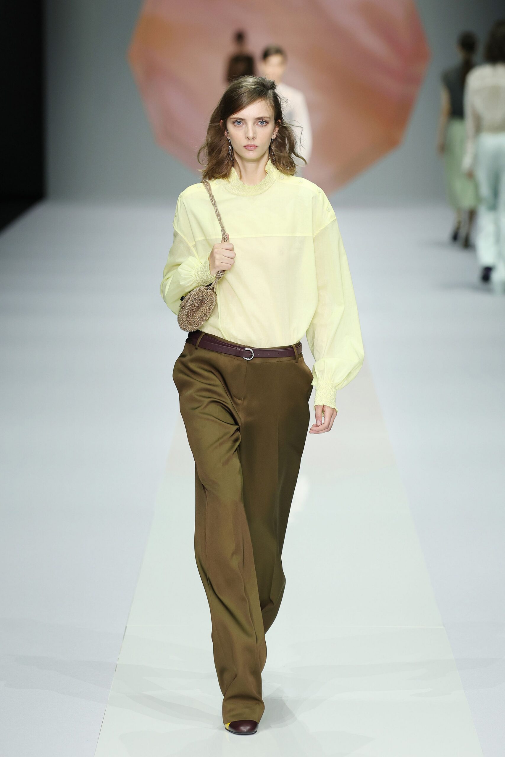 Butter-yellow blouse with a smocked collar and gathered cuffs. Small round raffia bag on a rope-thin strap. Burgundy leather belt with a narrow D-ring buckle. Olive draped trousers breaking long over the shoe. Two-tone pumps, yellow vamps capped in oxblood. Walking to lunch through a conservatory after the rain lets up.
