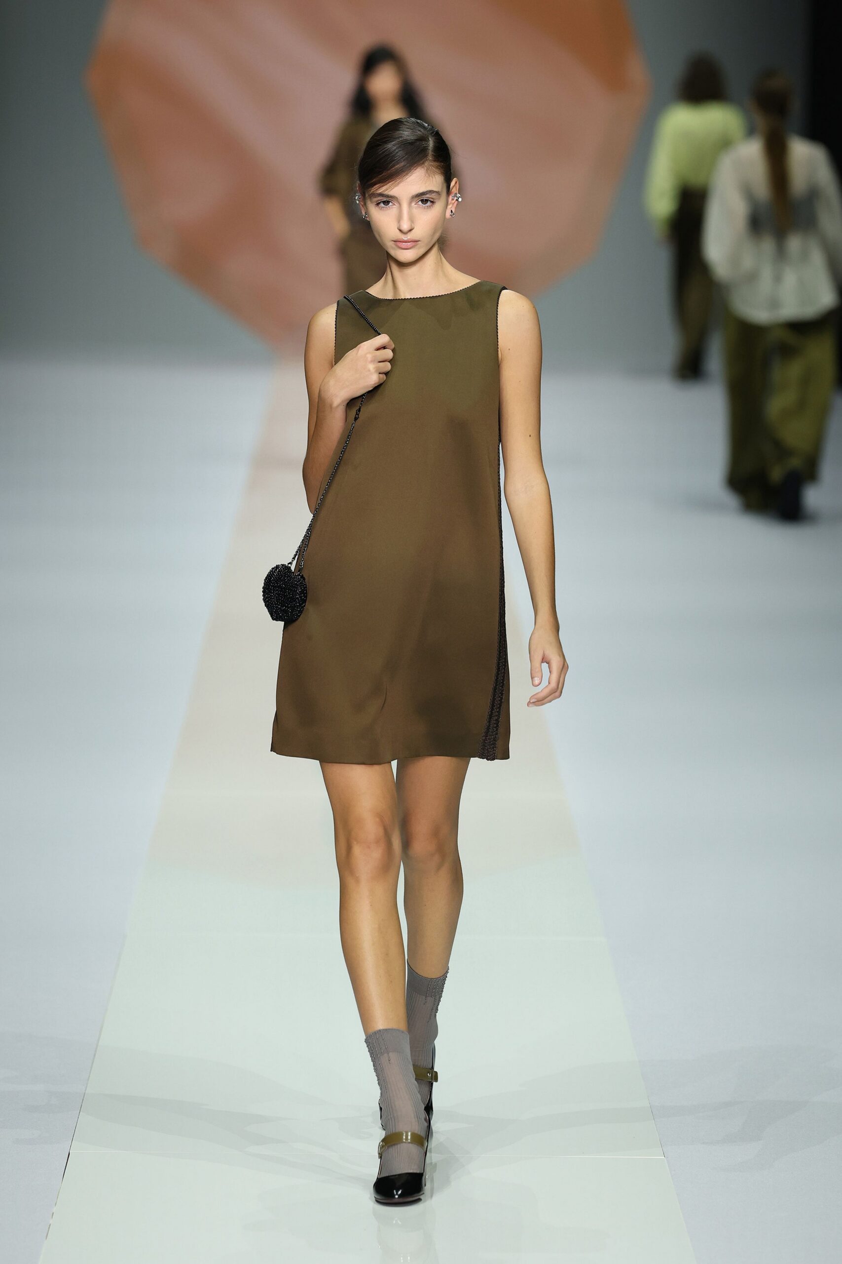 Olive shift dress in matte satin, black beaded trim running the side seams. Small black beaded sphere on a fine shoulder chain. Sheer grey ribbed ankle socks. Black patent cap-toe heels with an olive strap across the instep. Walking back from a museum lunch with a plum in your bag.