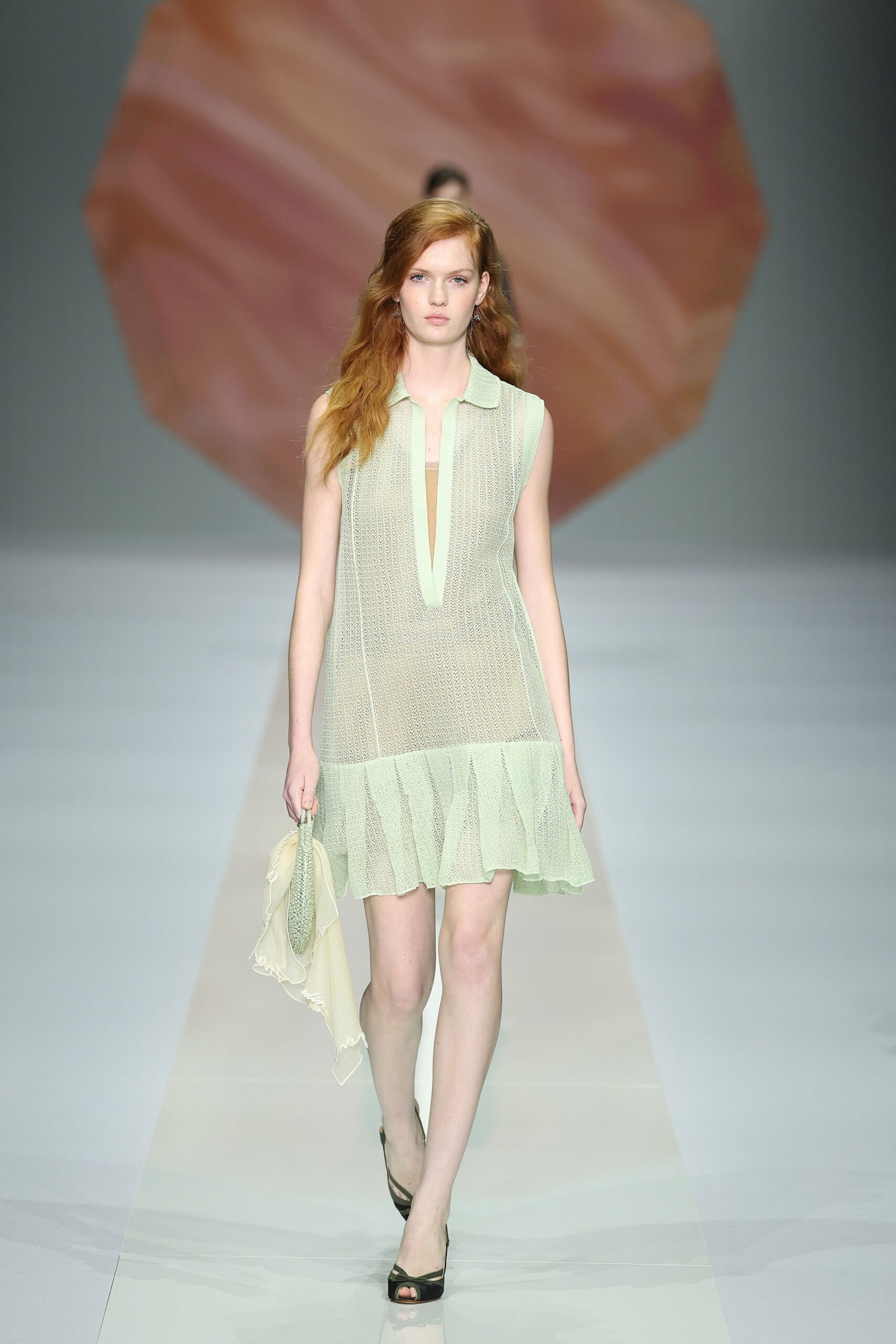 Pale pistachio open-knit dress with a plunging polo placket and flounced hem. Matching knit pouch wrapped in a sheer scarf, loose threads trailing. Black peep-toe heels with narrow crossover straps. Walking home from a beach wedding after the light has gone milk-blue.