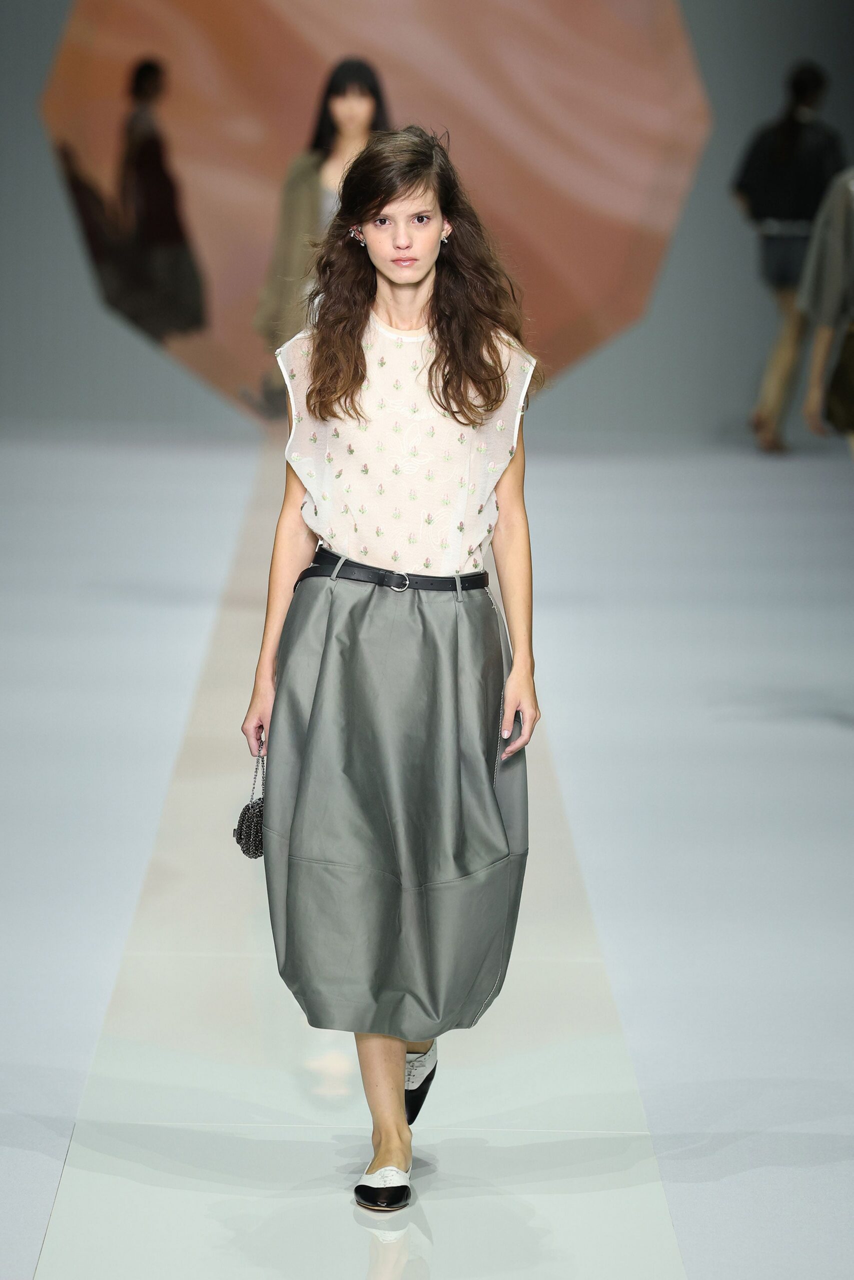 Sheer ivory shell top sprinkled with tiny pastel sprig embroidery. Narrow black leather belt with a small oval buckle. Dove-grey skirt cut in stiff panels, ballooning into a rounded hem. Black beaded pouch on a fine chain strap. White flats with glossy black cap toes. Walking back from a florist with stems wrapped in damp paper.