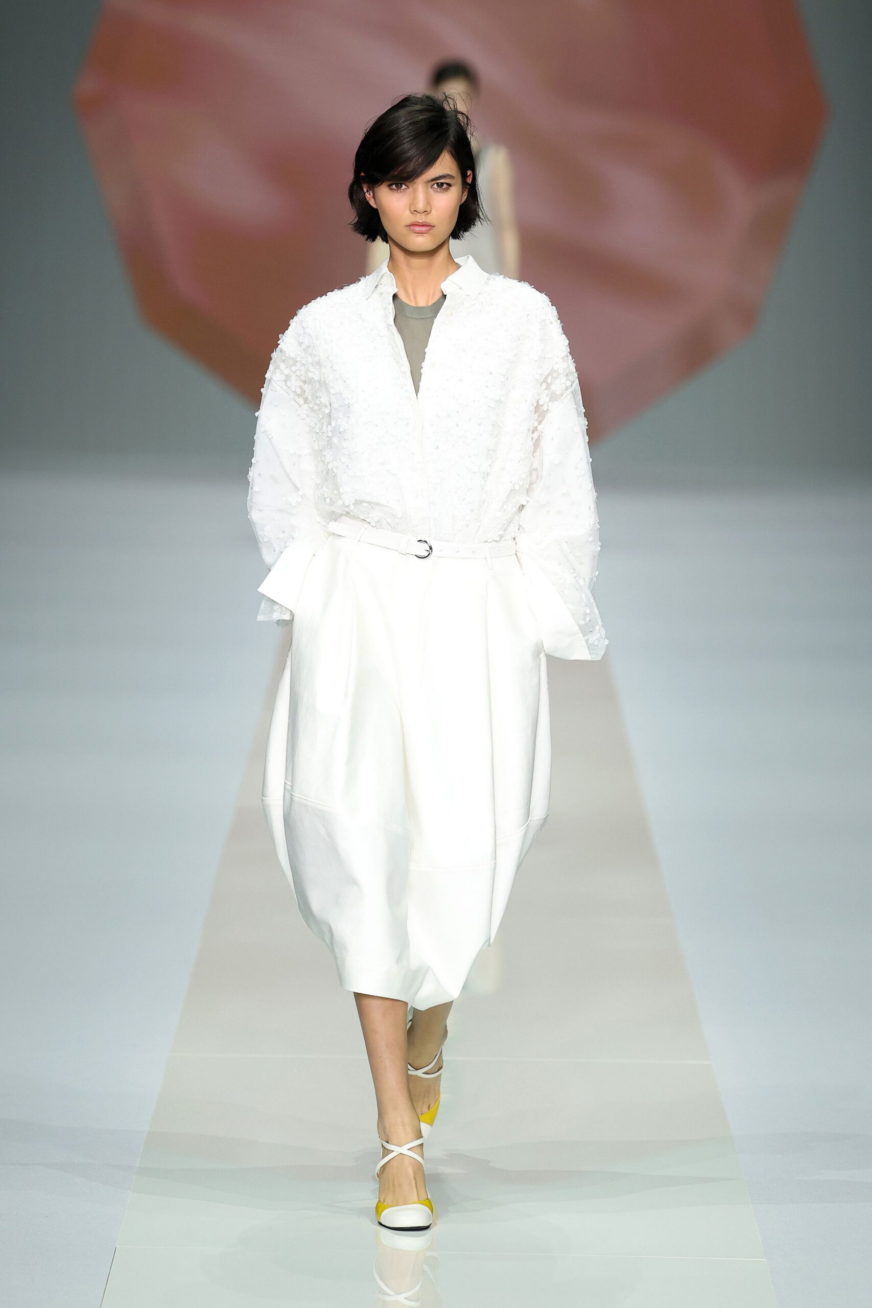 White blouse dusted with clipped floral tufts, collar standing slightly open. Pale sage underlayer peeking cleanly at the V neck. White cocoon skirt in smooth bonded cloth, cinched by a slim D-ring belt. White crisscross flats with citron toe caps. Museum atrium at noon, carrying lilies still wrapped in paper.