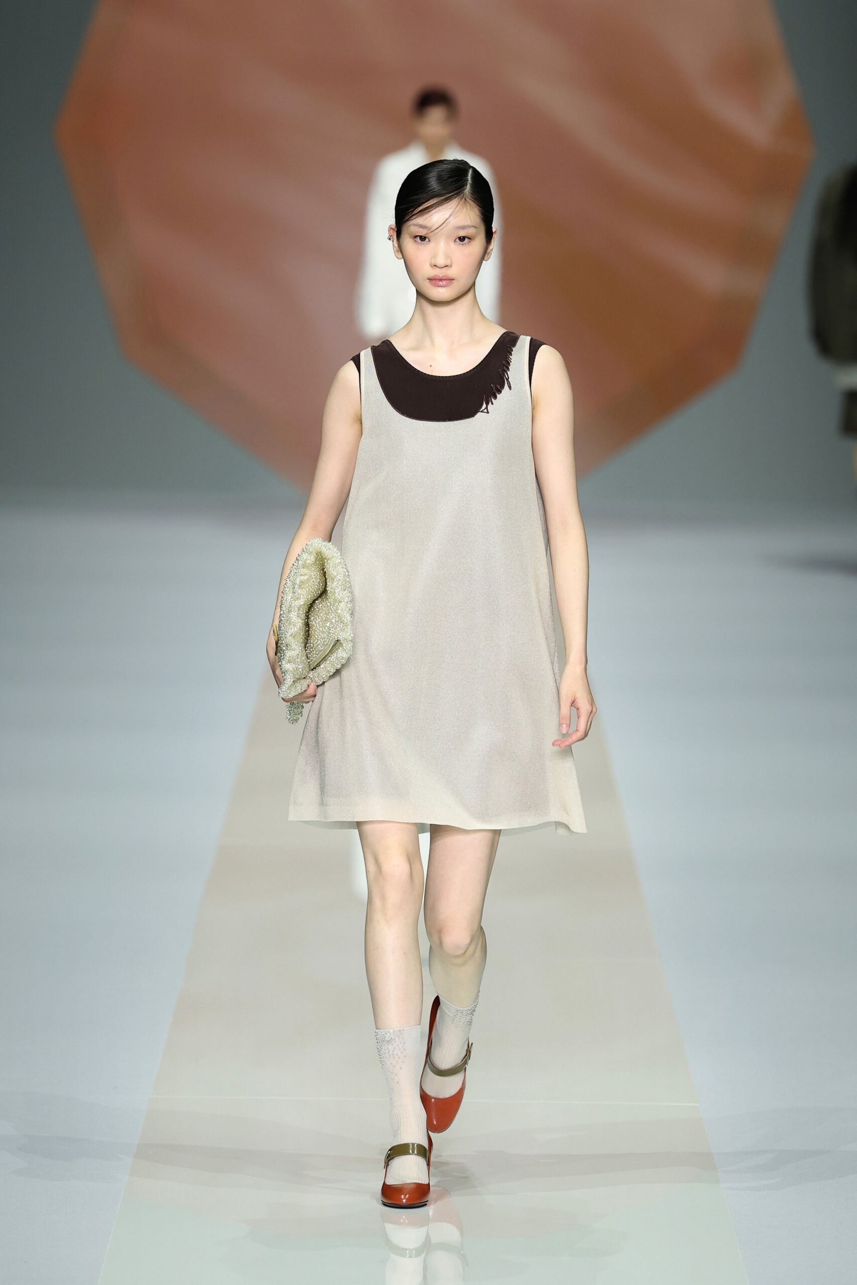 Pale greige A-line shift with a softly brushed surface. Dark brown underlayer, one shoulder edge left in loose fringe. Slouchy bouclé clutch tucked under the arm. Cream openwork ankle socks. Burnt orange patent Mary Janes with an olive instep strap. Crossing a museum atrium before the café opens and the floor still echoes.