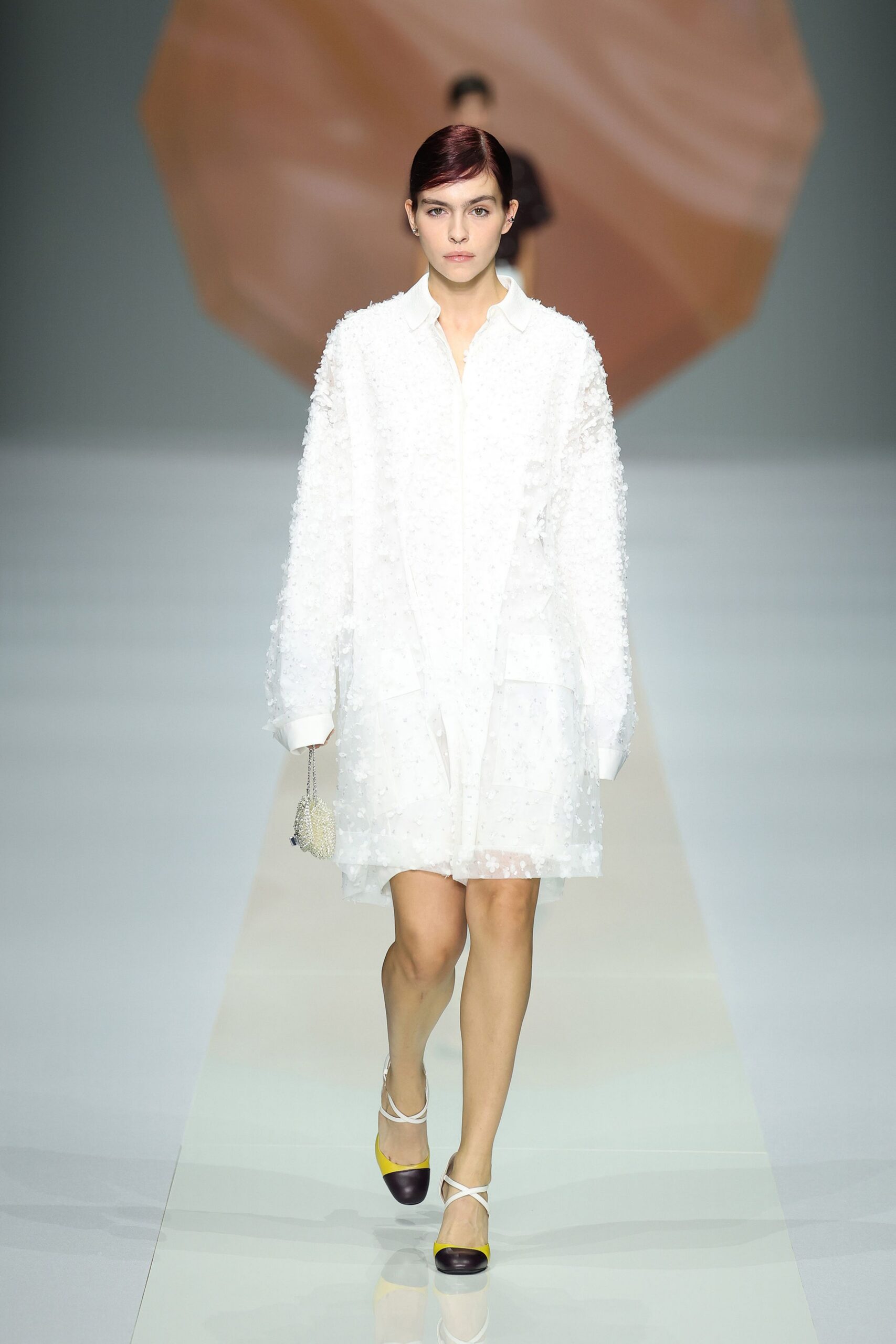 White sheer organza shirt dress scattered with tiny appliqued blossoms. Beaded drawstring pouch hanging from a thin silver chain. White strap heels with lemon vamps and deep aubergine toe caps. Crossing a hotel atrium after somebody else's wedding breakfast.