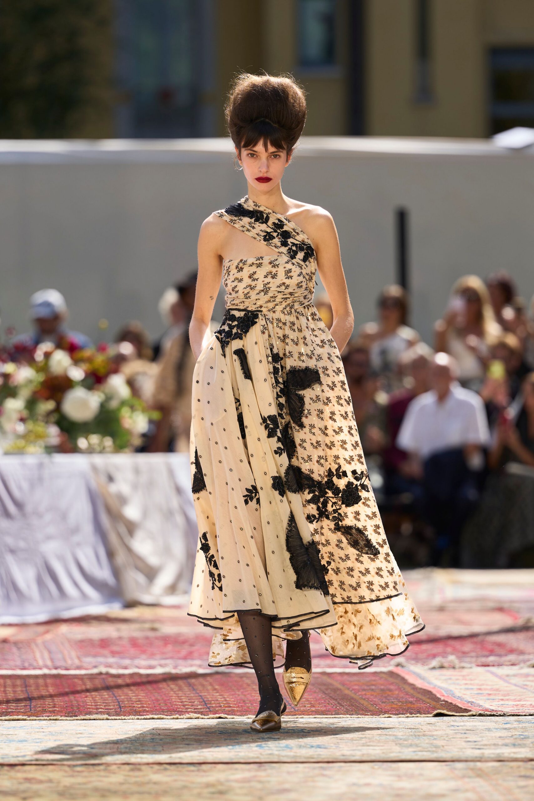 Towering beehive updo with a dark underfringe cut straight across. One-shoulder cream tulle dress with black floral appliqué and a draped side panel. Black sheer tights pricked with tiny dotted crystals. Gold pointed flats with a gathered vamp. Garden luncheon running late, rugs laid across the grass.