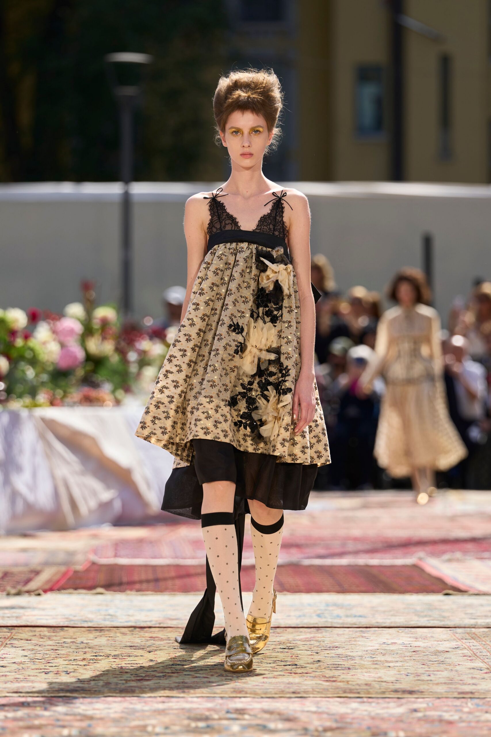 Empire dress in cream floral jacquard, black lace cups tied with tiny shoulder bows. Oversized organza flowers climbing one side over black floral lace. One ivory dotted knee sock, one sheer black stocking. Gold loafers with a creased metallic finish. Garden luncheon running late, sunlight catching on the dessert spoons.