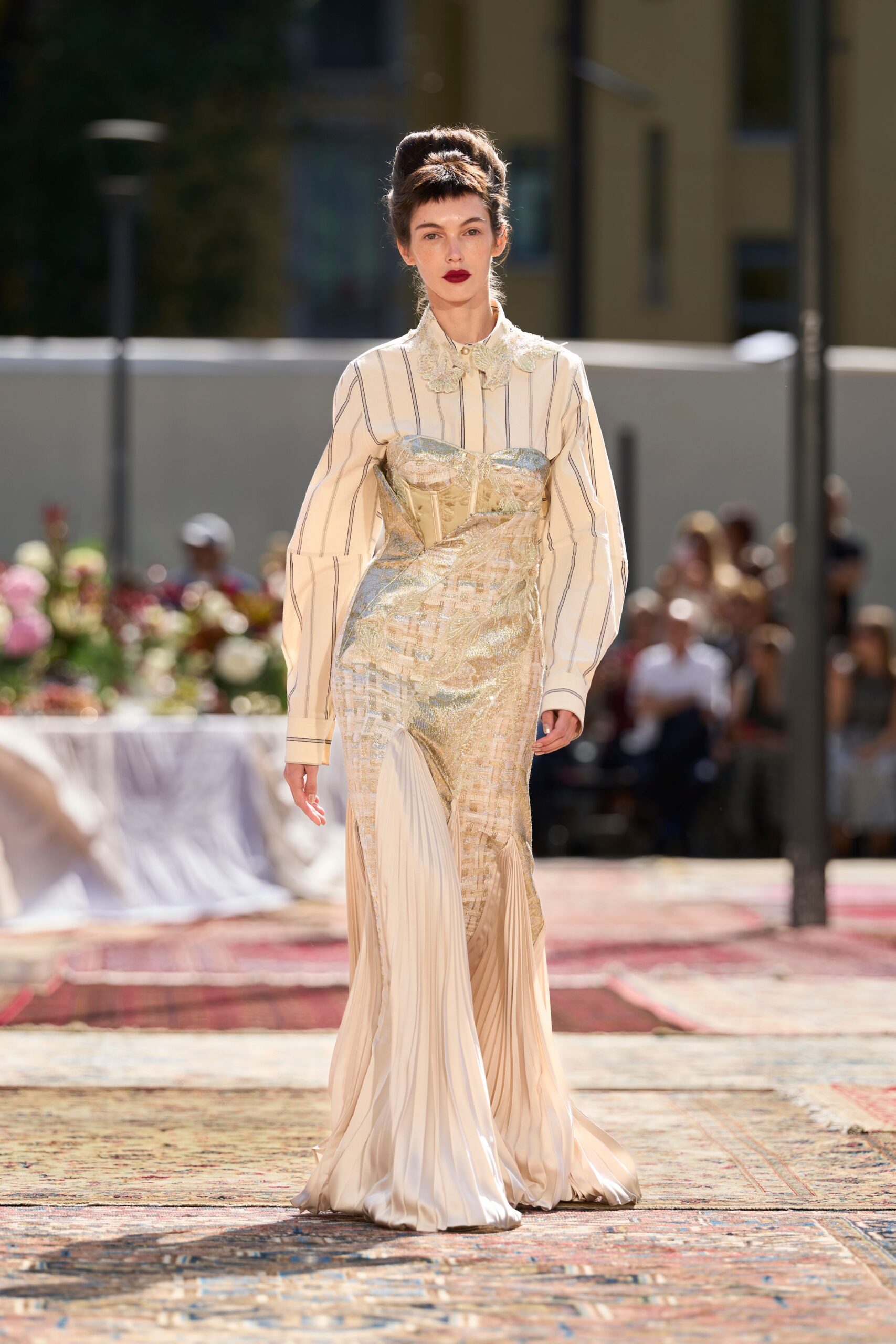 Cream striped shirt with balloon sleeves and a lace-edged Peter Pan collar. Pale metallic corset gown draped askew, split by ivory knife pleats at the hem. Garden wedding lunch gone long, sun bouncing off the cutlery.