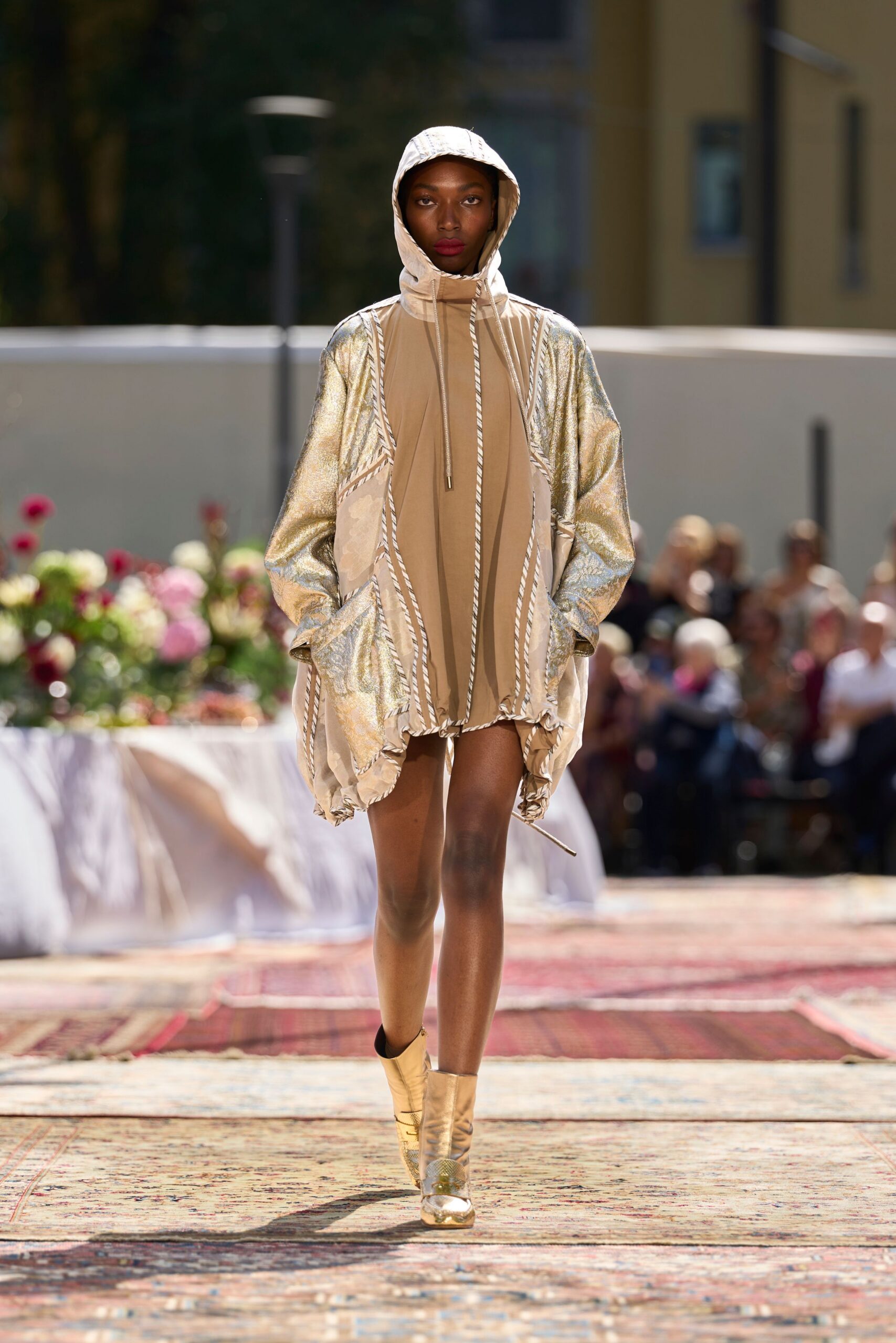 Sand nylon mini dress with a high zip neck and bubbled hem. Champagne metallic sleeves and cream panels piped in twisted rope trim. Gold ankle boots with mirrored toes and a mesh-textured cap. Crossing a hotel courtyard after sunset, still carrying the heat of the pool.