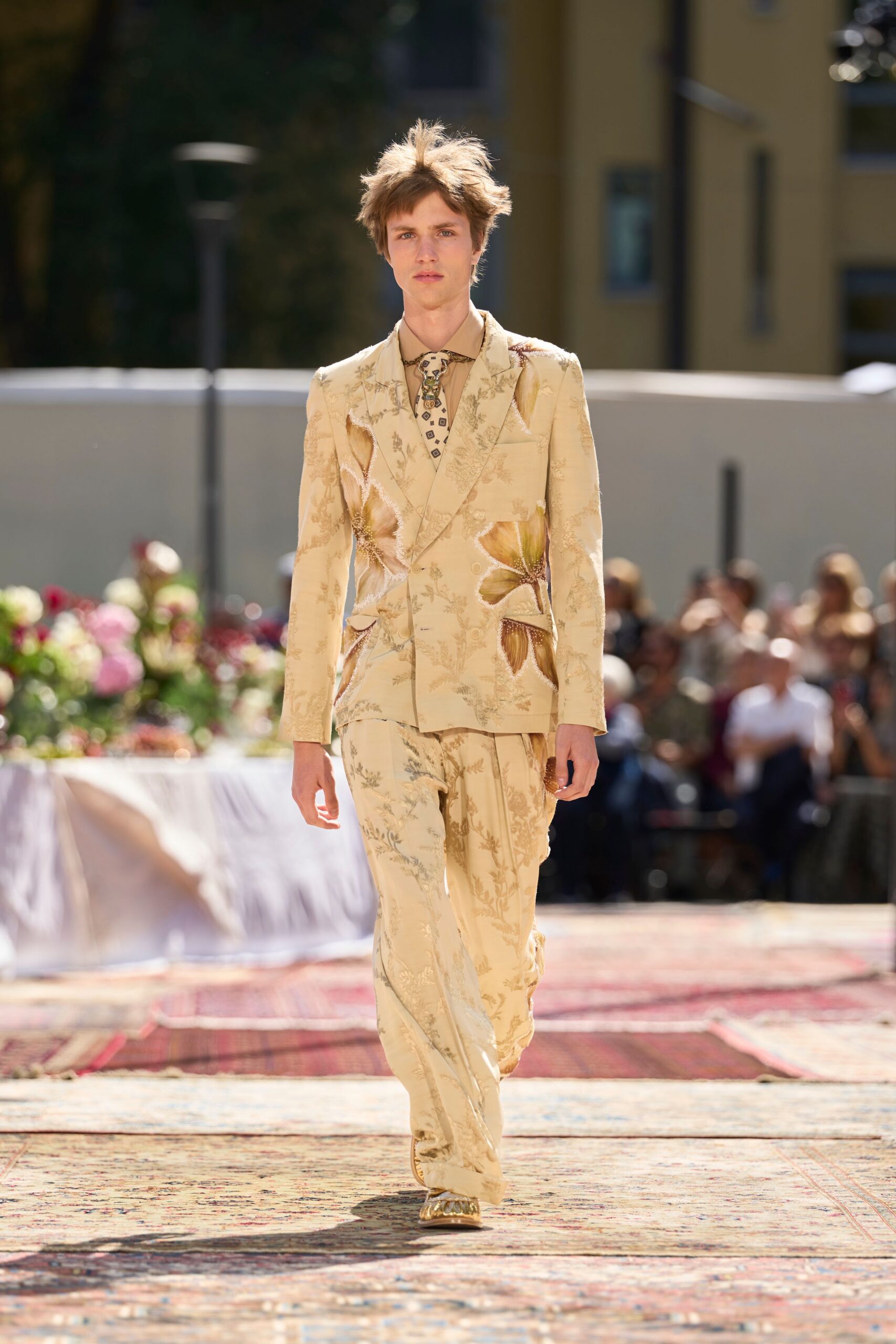 Sand shirt buttoned to the throat. Beaded bird-skull slide with a diamond-tipped hanging strap. Buttercream floral brocade jacket, raw-edged cutouts opening into oversized blooms. Matching brocade trousers breaking softly over the shoe. Gold ruched loafers. Walking across a hotel courtyard after someone else's garden wedding.