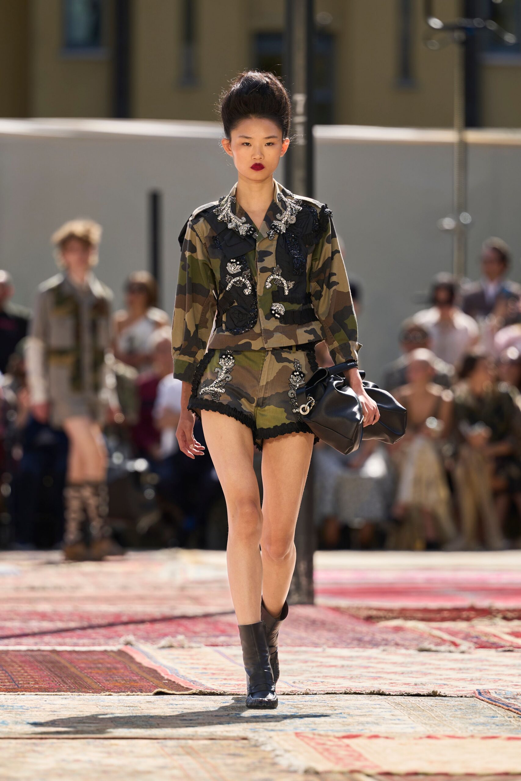 Cropped camouflage jacket crusted with silver beading and black ruffled trim. Matching camo shorts, crystal sprigs climbing the fly, hem softly frayed. Slouchy black leather carryall with oversized silver grommets. Black ankle boots with ribbed rubber toes. Crossing a hotel courtyard after noon, still carrying last night's bag.