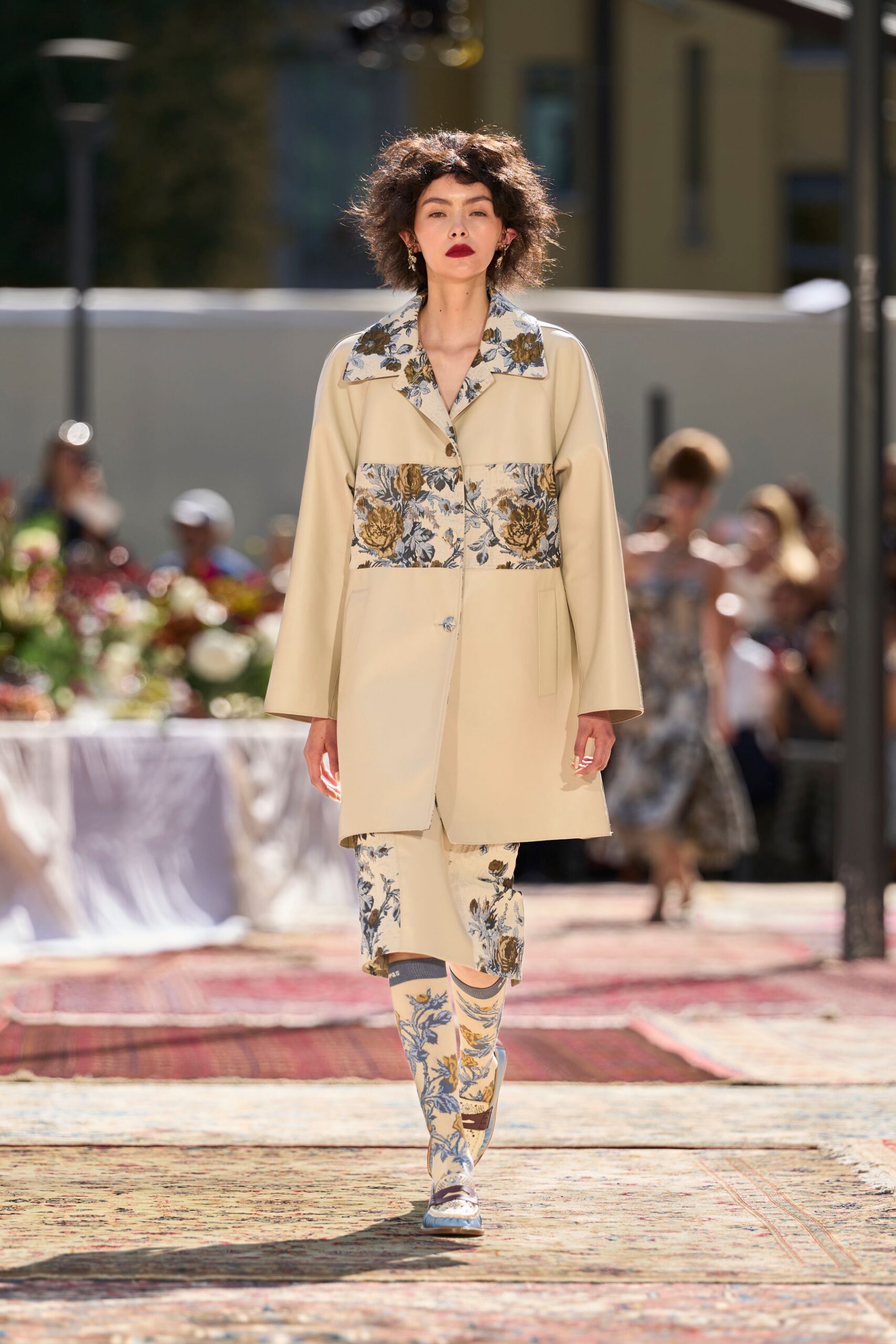 Cream coat with wide floral brocade collar and matching chest panel. Cream pencil skirt cut in the same blue-and-ochre floral weave. Knee socks printed to match, grey bands gripping the calves. Two-tone loafers with a square apron seam and brown strap. Garden lunch running late, rugs laid over the paving stones.