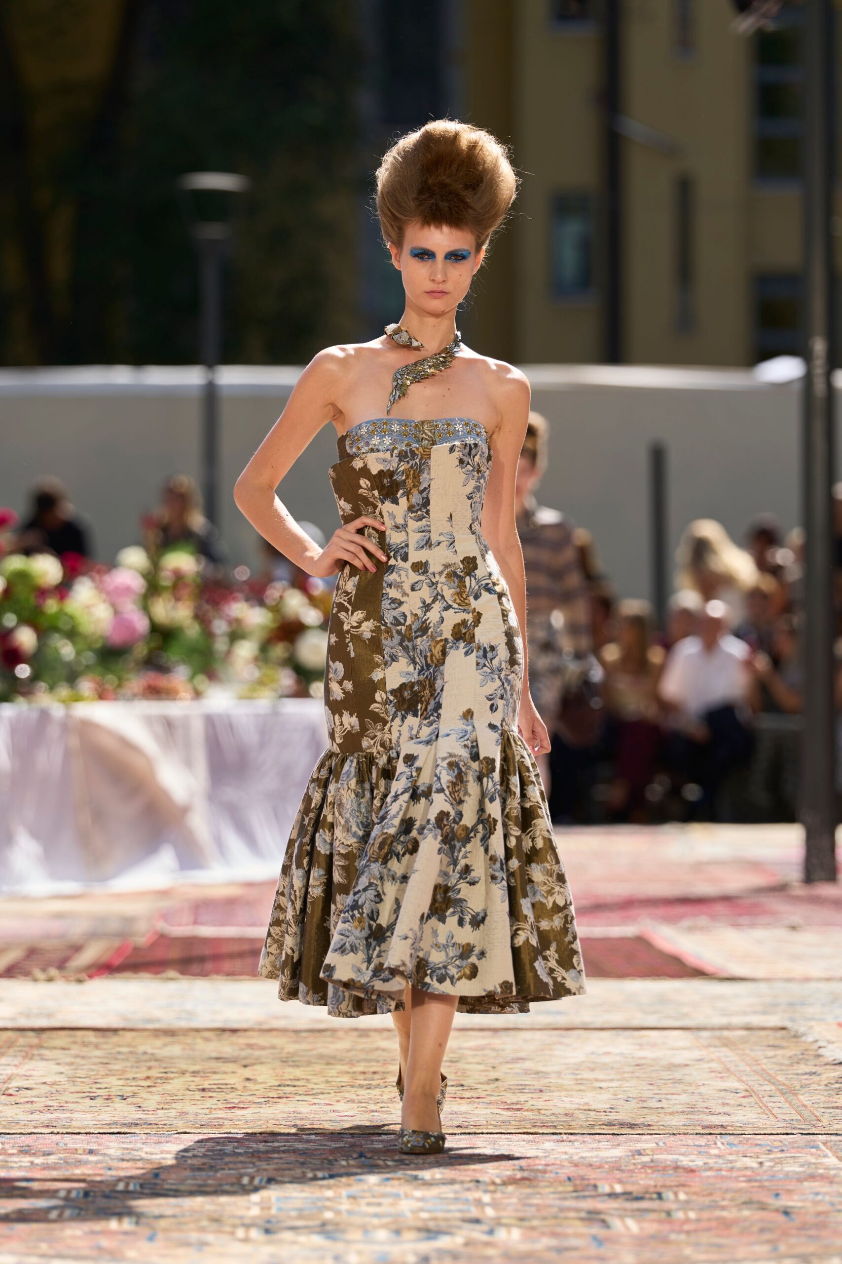 Asymmetric bird necklace curling round the throat in mottled metal. Strapless floral jacquard dress pieced in brown, cream and powder blue panels. Matching brocade pumps with a narrow pointed toe. Garden luncheon set after the plates have been cleared for coffee.