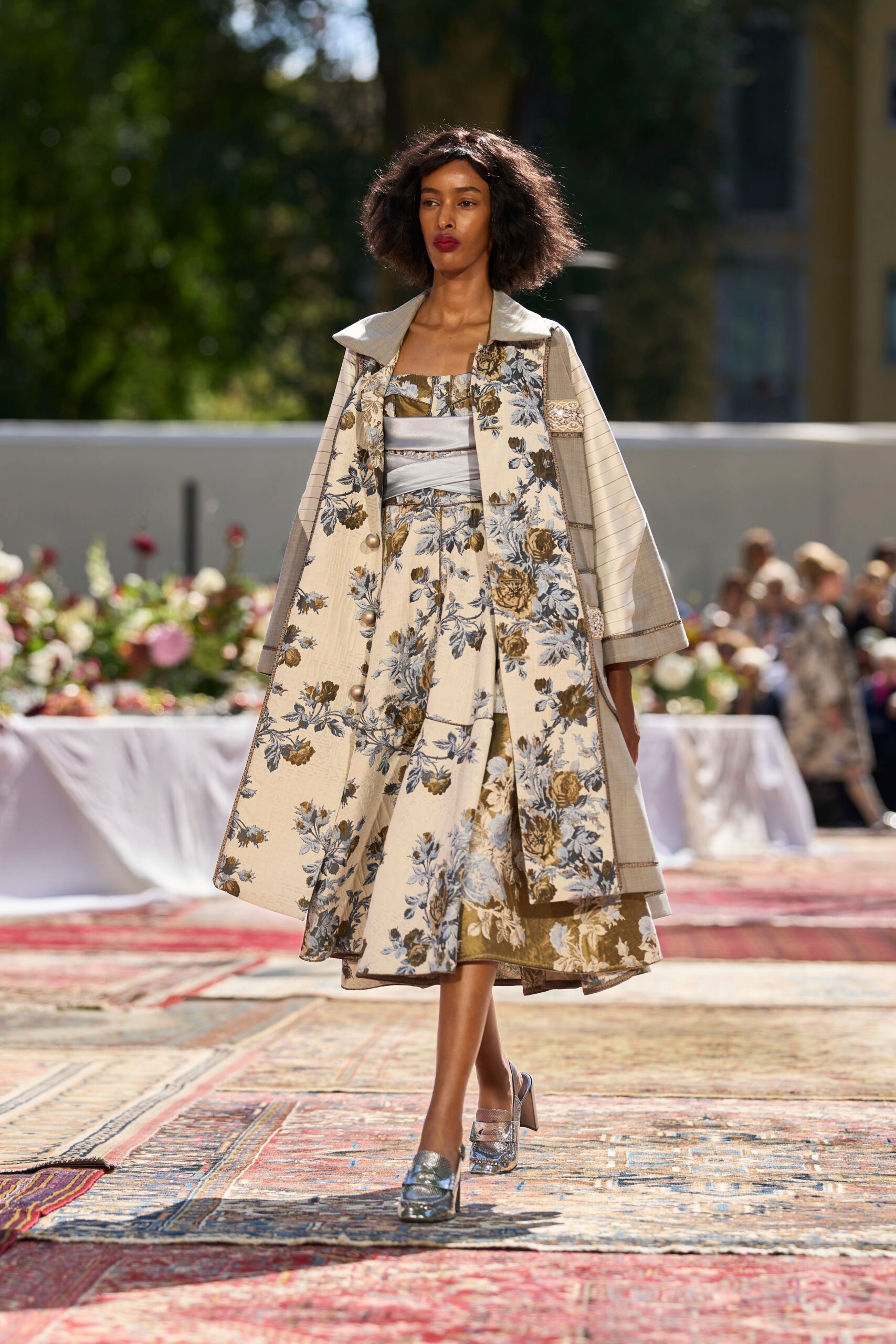 Cream floral brocade coat with a broad stand collar and ball buttons. Pale satin bandeau wrapped across the waist in overlapping bands. Full floral skirt with an olive border peeking from the uneven hem. Silver slingback heels in cracked metallic leather. Garden luncheon running long, tablecloths lifting in the afternoon heat.
