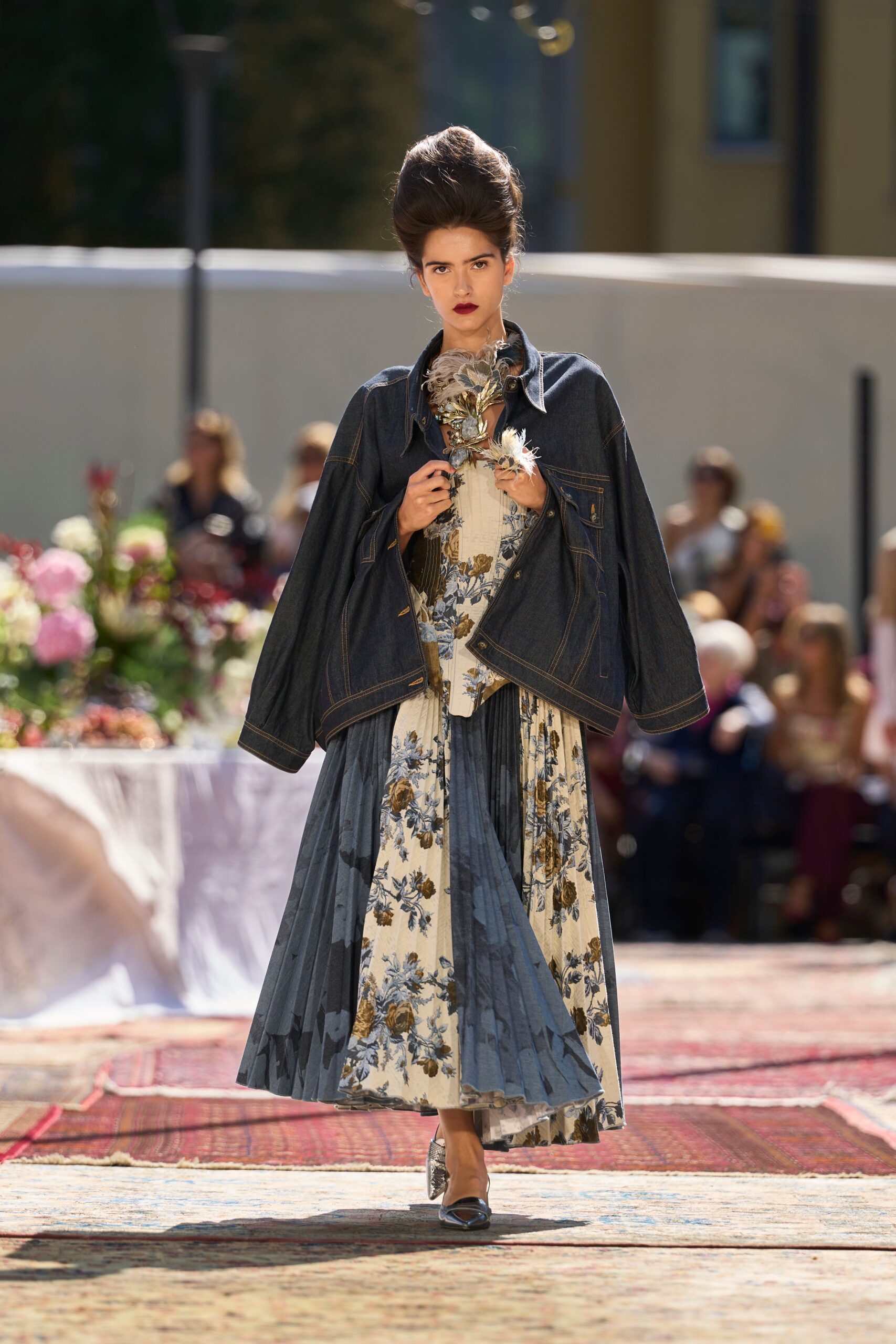 Metal leaves and pale feathers clustered high at the throat. Oversized raw-indigo denim jacket with tobacco topstitching and a low chest pocket. Cream floral dress with a corseted bodice and slate pleated panels. Silver pointed heels. Garden lunch running late, rugs laid across the grass.
