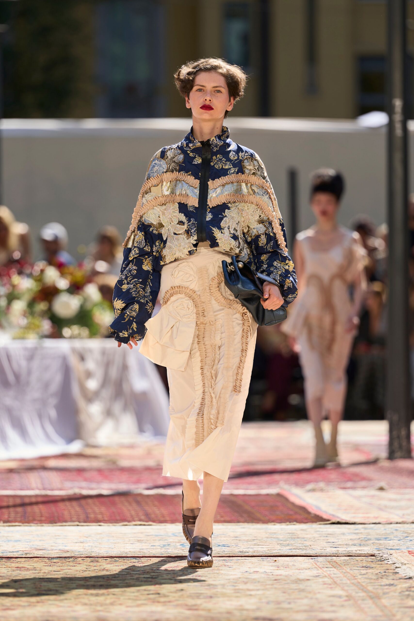 Navy floral brocade blouson with blush ruffle bands arcing across the chest. Ivory wrap skirt traced with ruched appliqué spirals and a bulky side flap. Slouchy black clutch tucked under one arm. Dark leather loafers with thick ridged soles. Crossing a hotel courtyard after lunch, still carrying the folded seating plan.