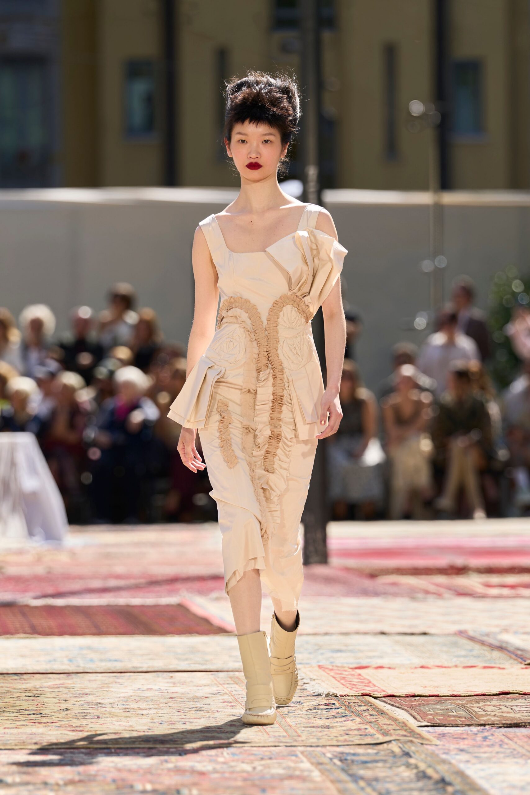 Cream dress with dense ruched channels and folded peplums jutting from hip and shoulder. Cream ankle boots with wrapped shafts and rounded molded toes. Crossing a tiled courtyard after a wedding lunch has run too long.