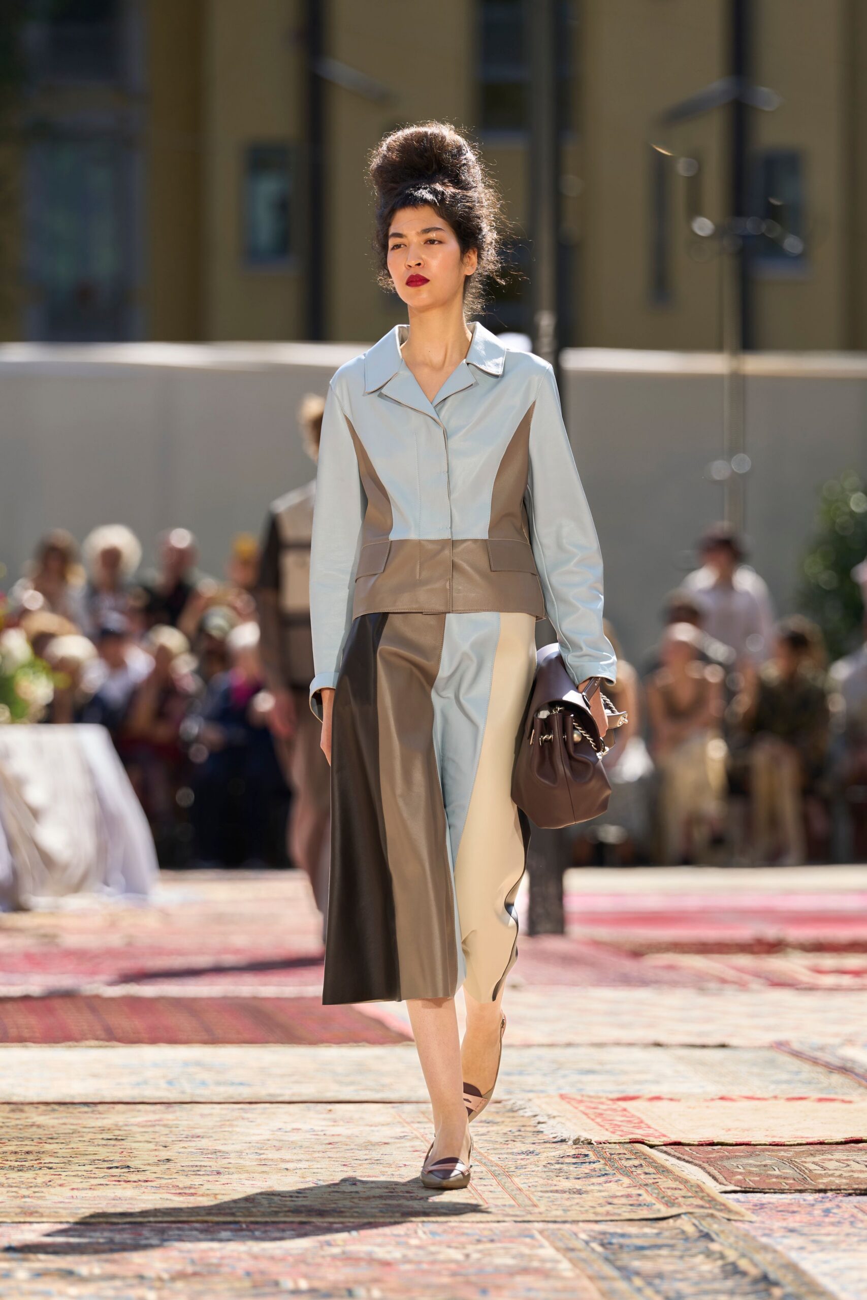 Powder-blue leather jacket, taupe side panels carving in the waist. Mismatched leather midi in cream, taupe and black vertical blocks. Chocolate drawstring bag with silver rings at the gathered top. Pointed flats with a dark cap toe. Crossing a tiled courtyard at noon with papers tucked under one arm.