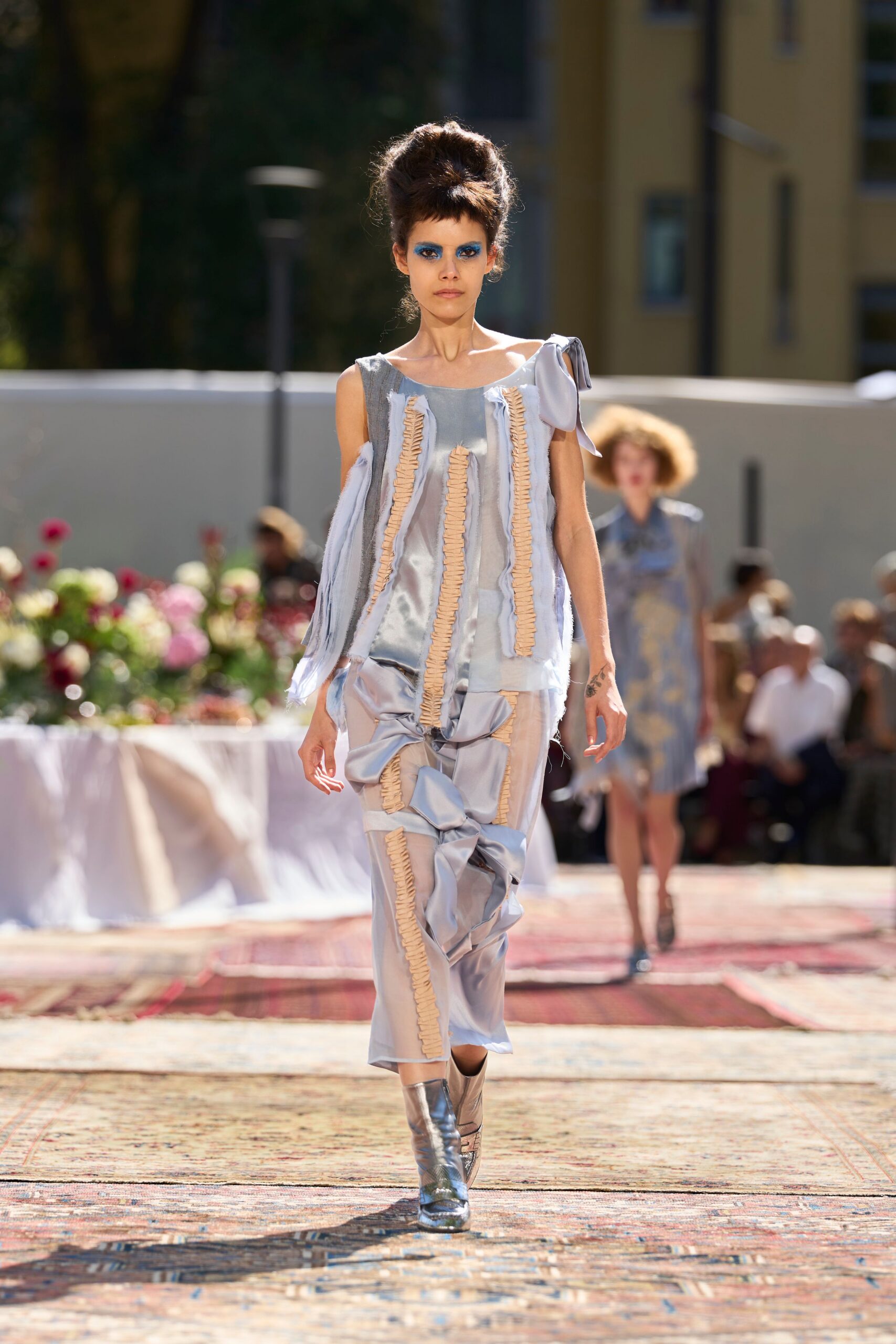 Ice-blue satin shift dress, raw strips and beige ruching running vertically. Matching satin trousers knotted into swollen bows down one leg. Silver ankle boots with mirrored shine. Garden lunch after the tablecloths are rumpled and the ice has melted.