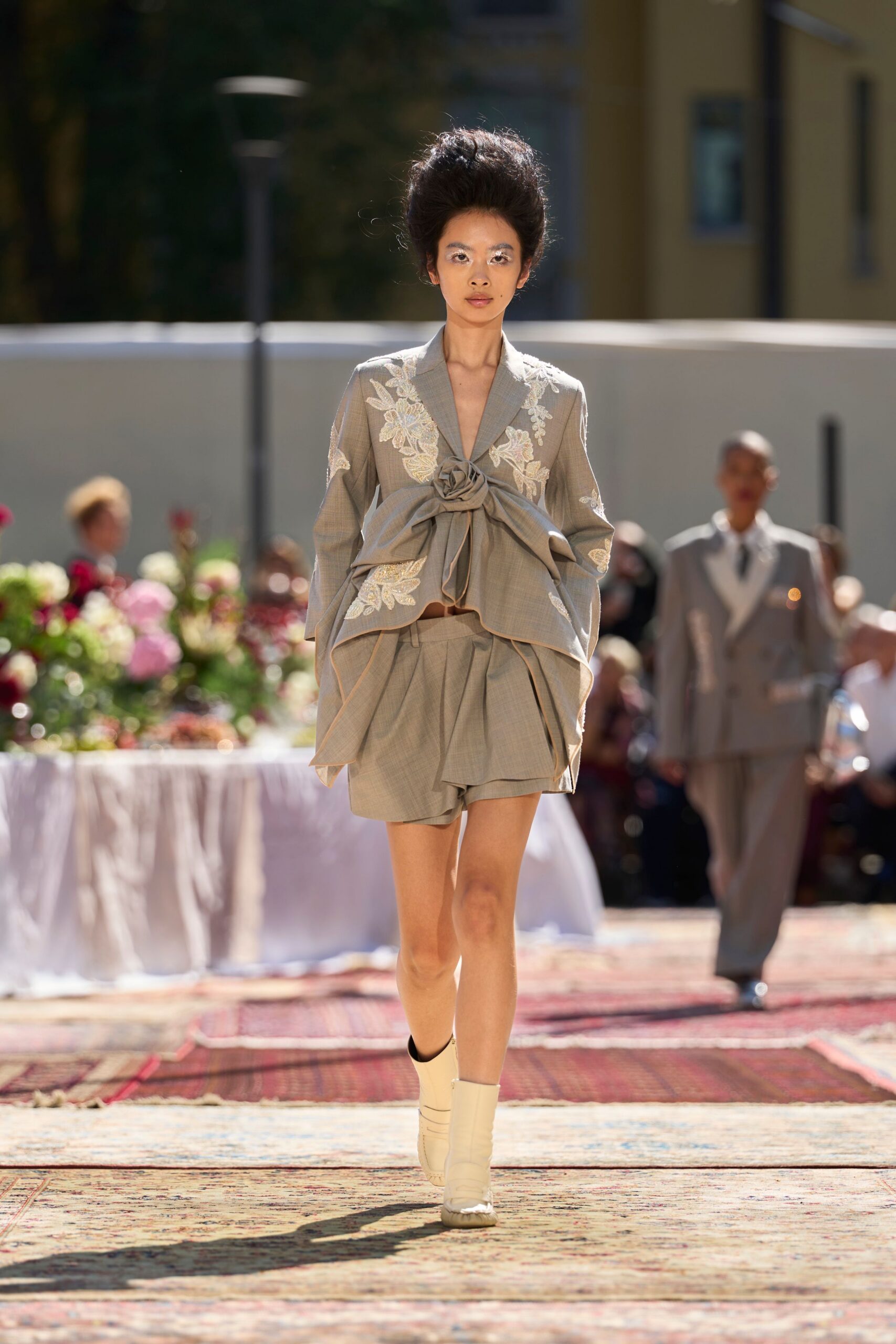 Taupe suiting jacket appliqued with ivory floral lace and tied in a giant rosette. Matching pleated wrap shorts, one panel hanging longer at the front. Cream ankle boots with split rubber soles. Garden lunch gone formal, heat rising off the paving stones.
