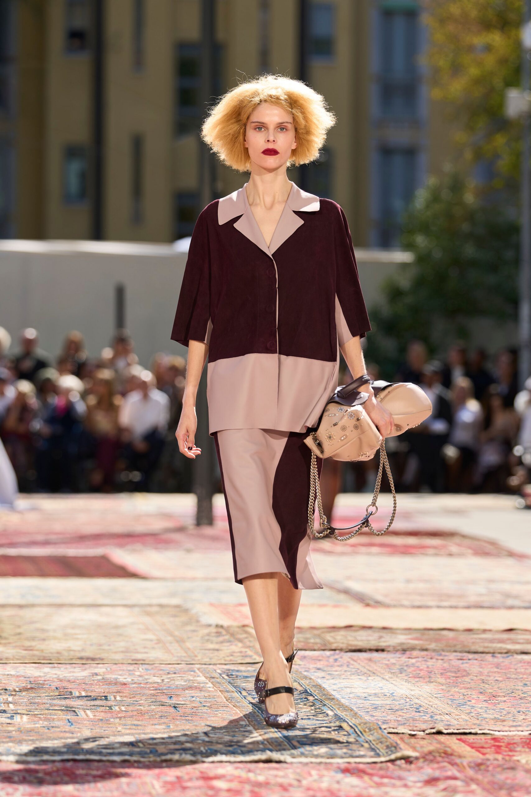Boxy blouse in oxblood and blush crepe, cut with broad lapels. Midi skirt in the same crepe, pieced into off-kilter color blocks. Blush leather duffel with crystal studs and drooping chain straps. Brown glitter Mary Janes with a narrow black instep strap. Crossing a hotel courtyard after a wedding breakfast ran long.